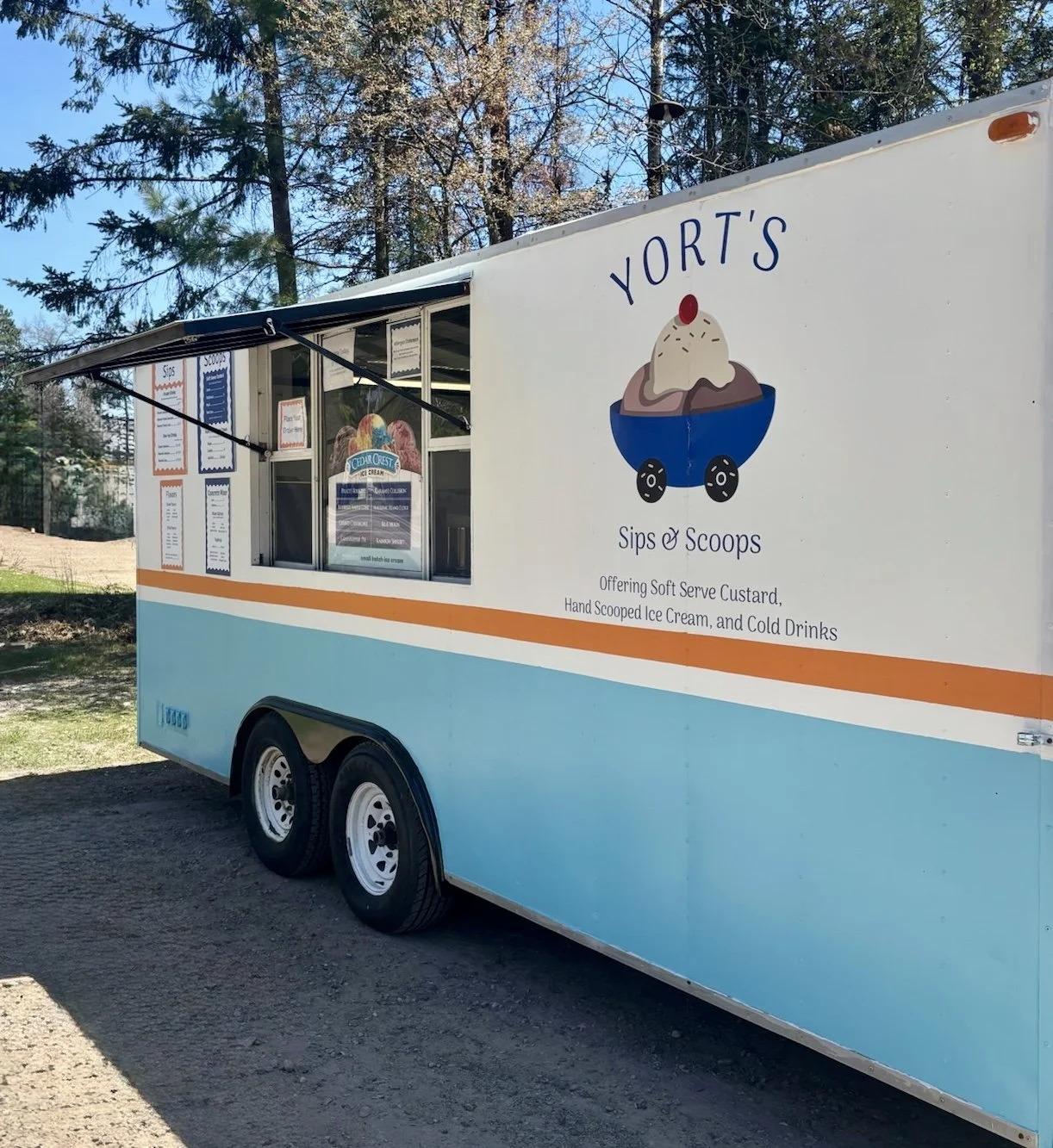 Ice cream truck called Yort's Sips & Scoops offering custard, ice cream, and cold drinks, parked outdoors with trees in the background.