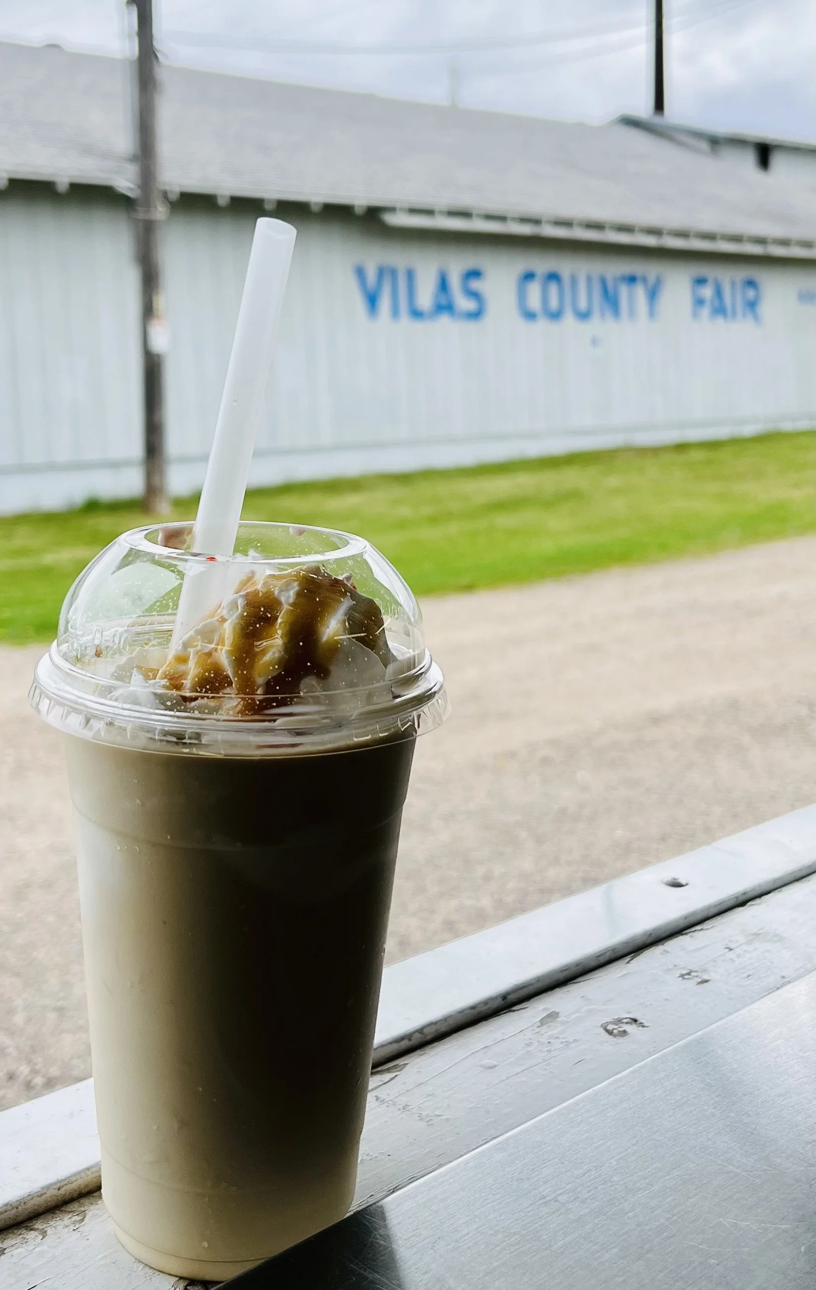 A cold beverage with caramel topping in a clear plastic cup with a straw, placed on a white wooden surface with the Vilas County Fair sign in the background.