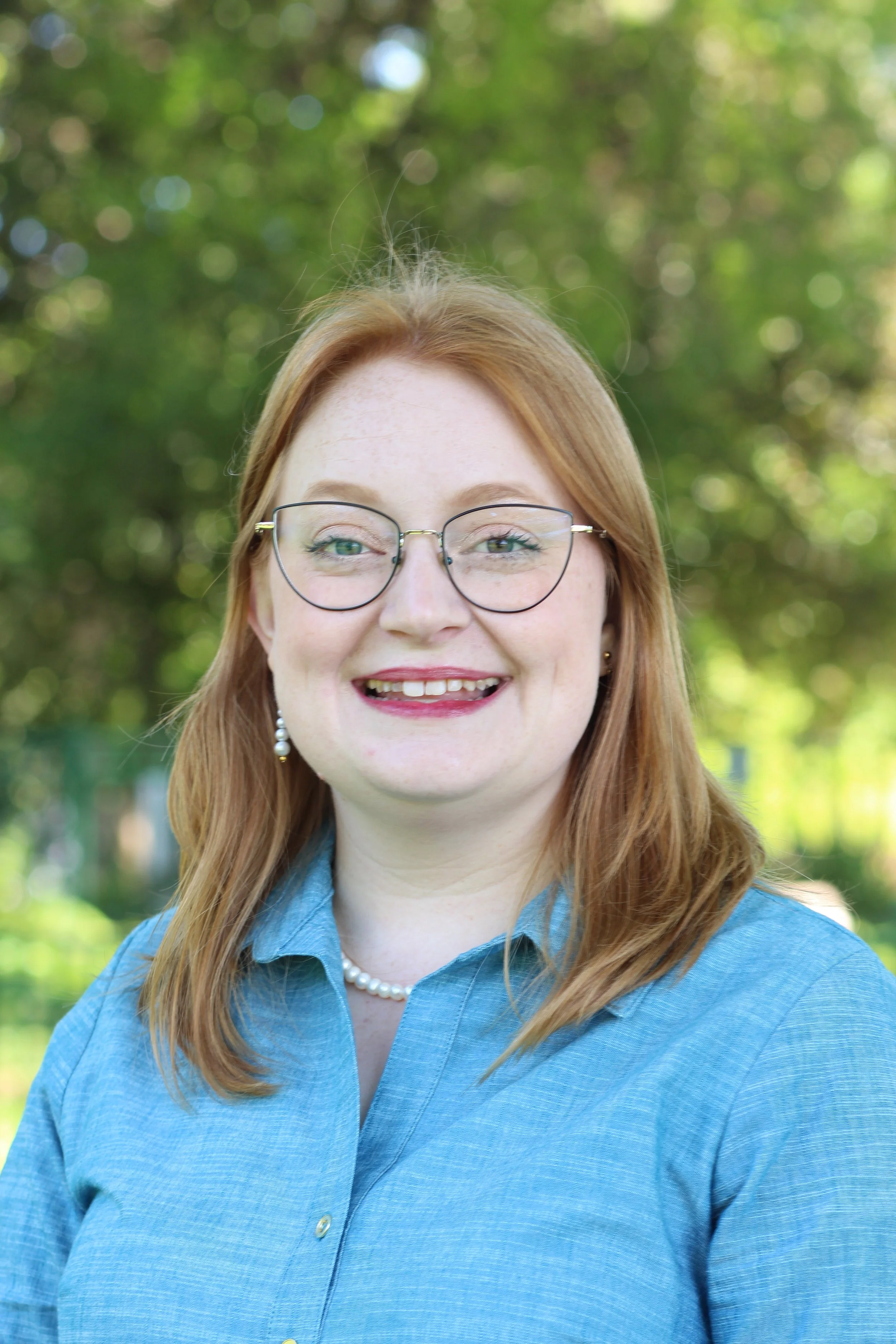 Vera Elwood, Candidate for Ward 2 City Council Person, A woman with red hair and green glasses is wearing a blue dress and standing in front of a line of trees. She is smiling at the camera.