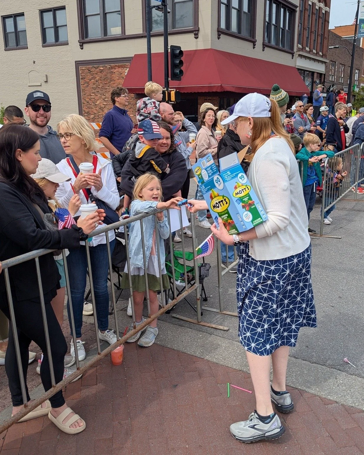 Vera Elwood, candidate for Ward 2 Council person in Columbia, Missouri, a redheaded woman in a blue dress and white cardigan hands fruit snacks to an excited young child on the side of a parade route