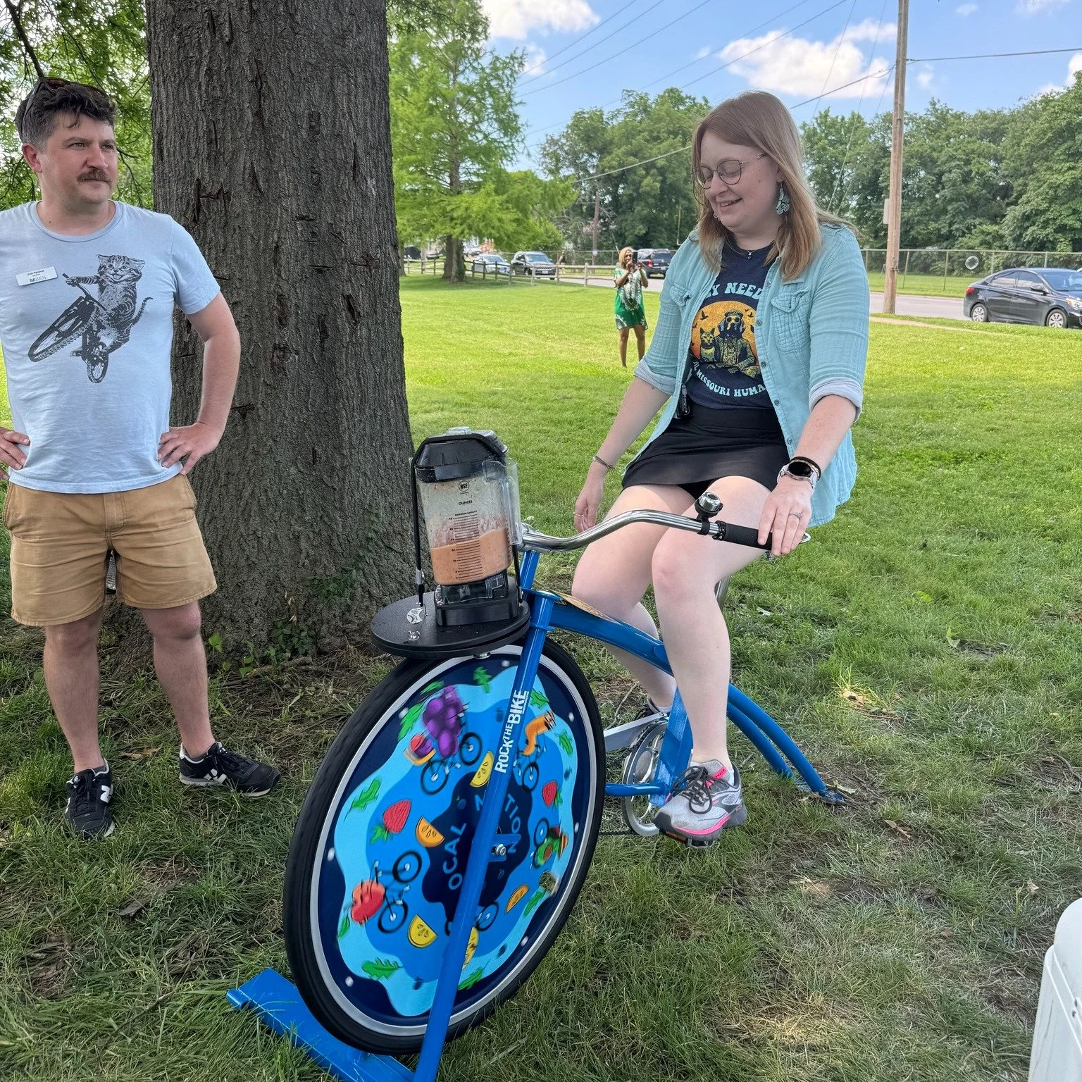 Vera Elwood for CoMo Ward 2 Council person, a  redheaded woman in a blue shirt and black skirt sits on a stationary bike. The bike is powering a blender to create smoothies.