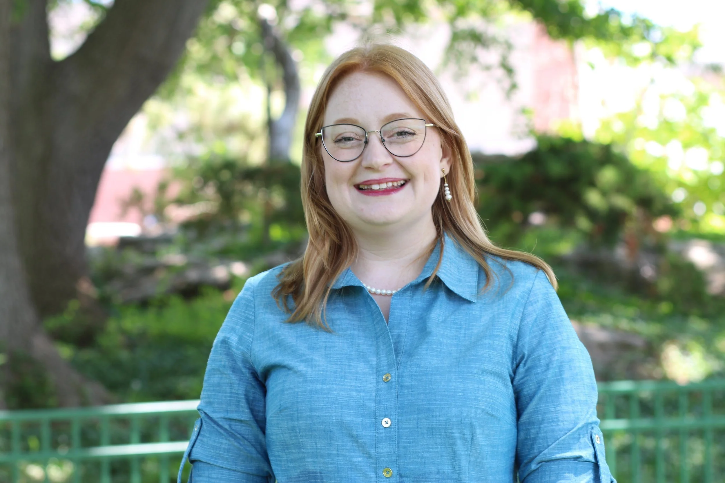 A woman with red hair and green glasses stands in front of a line of trees. She is wearing a blue button up dress with pearl earrings and is smiling at the camera.
