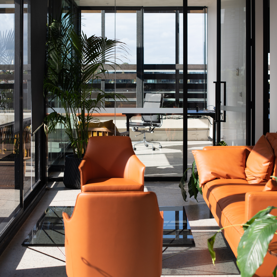 Modern office lounge area with orange chairs and a sofa, glass coffee table, and large potted plant, adjacent to a glass-walled office with a desk and chair, with city view outside.