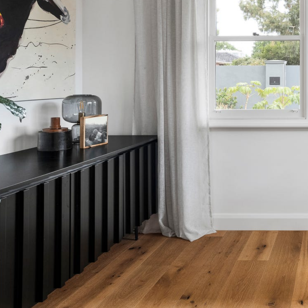 Part of a modern living room showing a black sideboard against a white wall, decorated with glass and ceramic vases, a framed black-and-white photograph, and abstract wall art. There is a window with white curtains and wooden flooring.