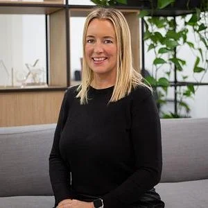 A woman with blonde hair, wearing a black long-sleeve top, sitting on a gray couch in a modern indoor setting with large windows and green plants in the background.