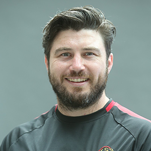 A man with dark hair and a beard smiling, wearing a dark sports shirt with red accents, against a plain background.