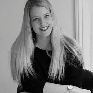 Black and white photo of a smiling woman with long blonde hair, wearing a black top, sitting by a window.