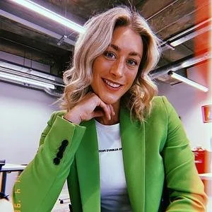 A woman with blonde hair smiling and wearing a bright green blazer and a white top in an industrial-style office or studio setting.