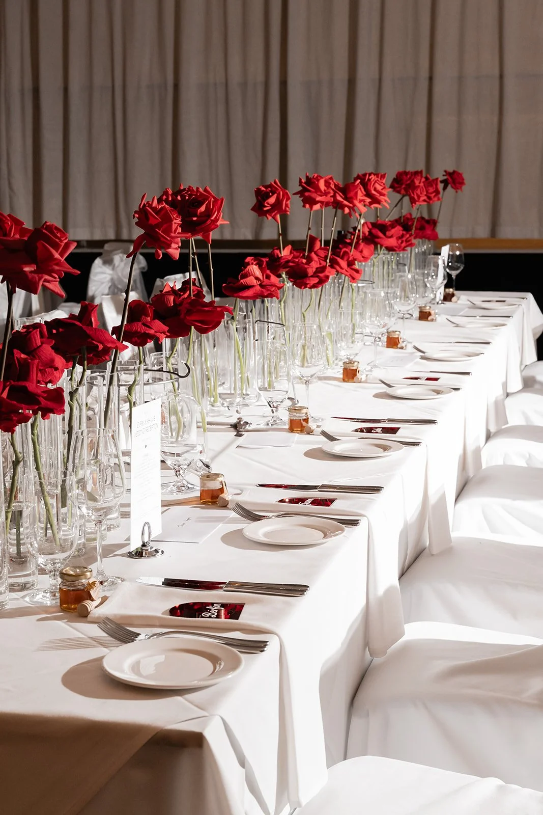 Elegant banquet table set with white tablecloths, tall red floral centerpieces, glassware, white plates, and silverware.