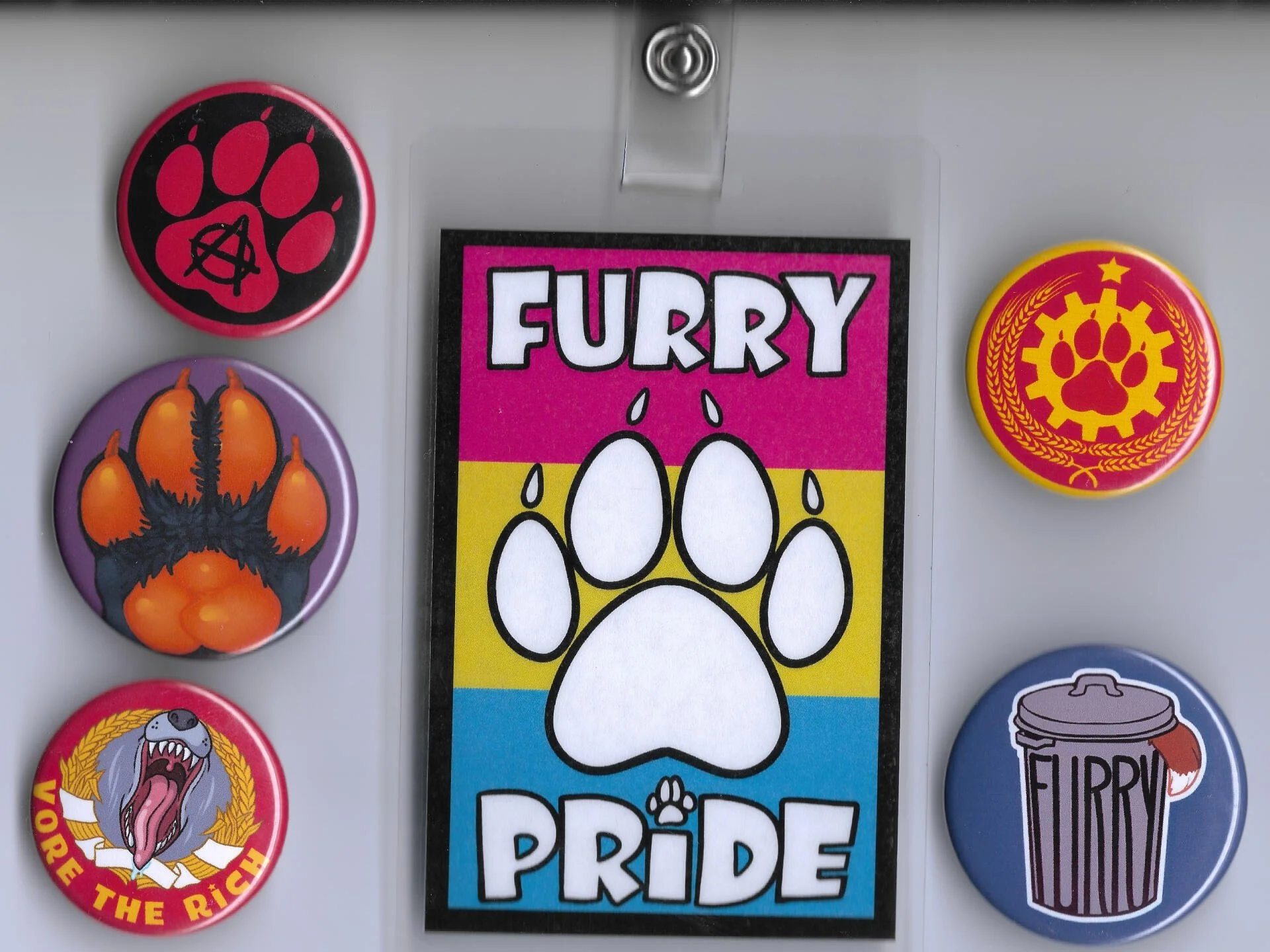 Five colorful pins and a card with a large paw print design. The pins include a pink and black 'furry' pin with a paw and anarchy symbol, a purple and orange pin with a cartoonish creature, a yellow and red pin with a paw and gear design, a yellow and red pin with a wolf head and the phrase 'Vore the Rich,' and a blue and gray pin with a coffee cup and the word 'Furry'.