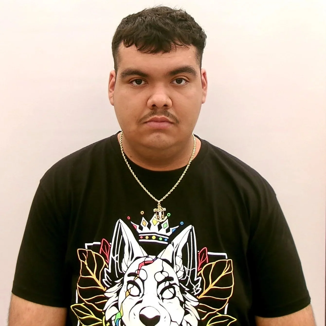 A young man with short dark hair and light skin, wearing a black T-shirt with a colorful graphic of a wolf's face, a crown, and leaves, along with a silver chain necklace with a pendant.