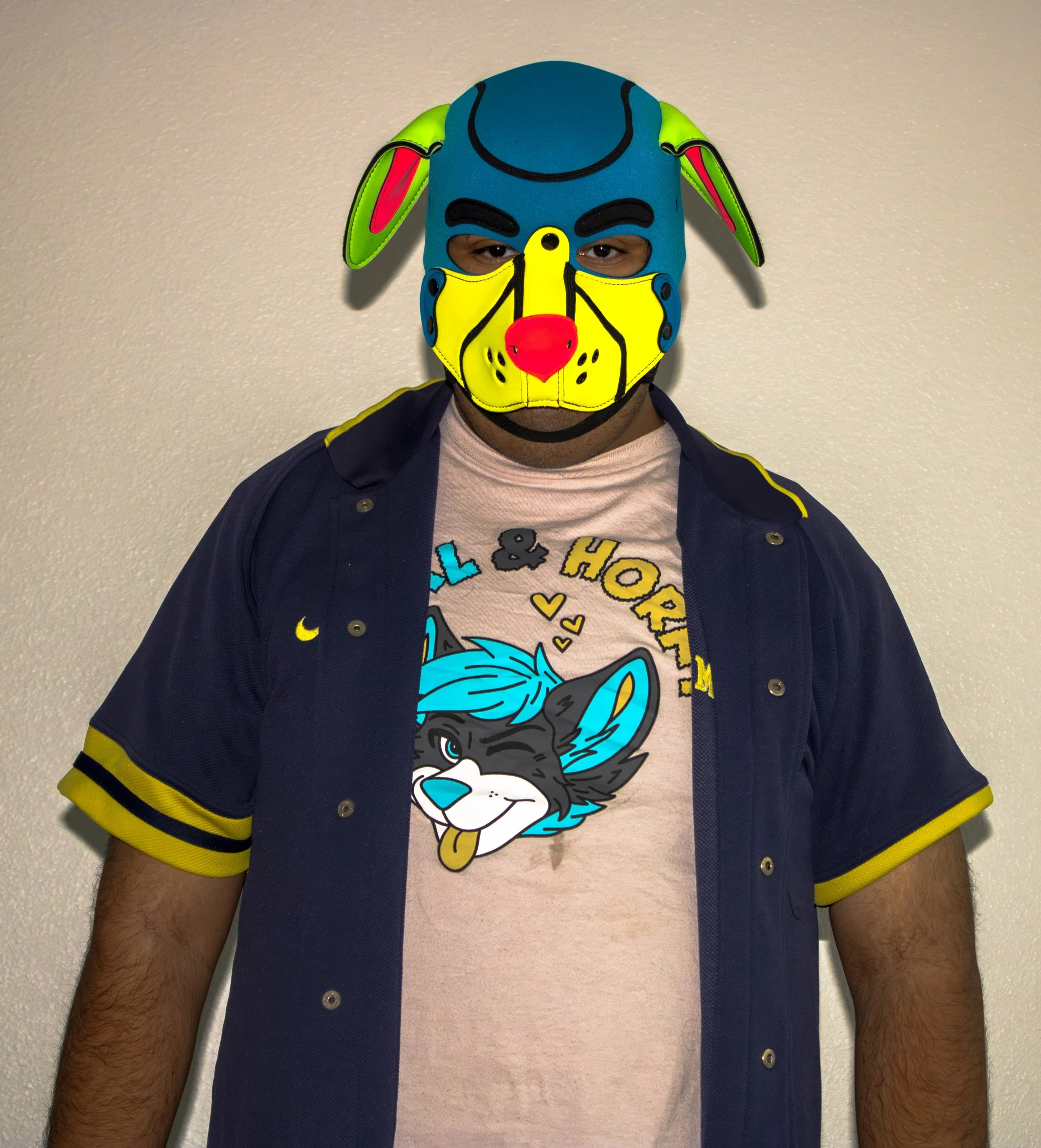 Person wearing a colorful cartoon dog mask, a navy blue jacket with yellow trim, and a light pink T-shirt with colorful text, standing against a beige wall.