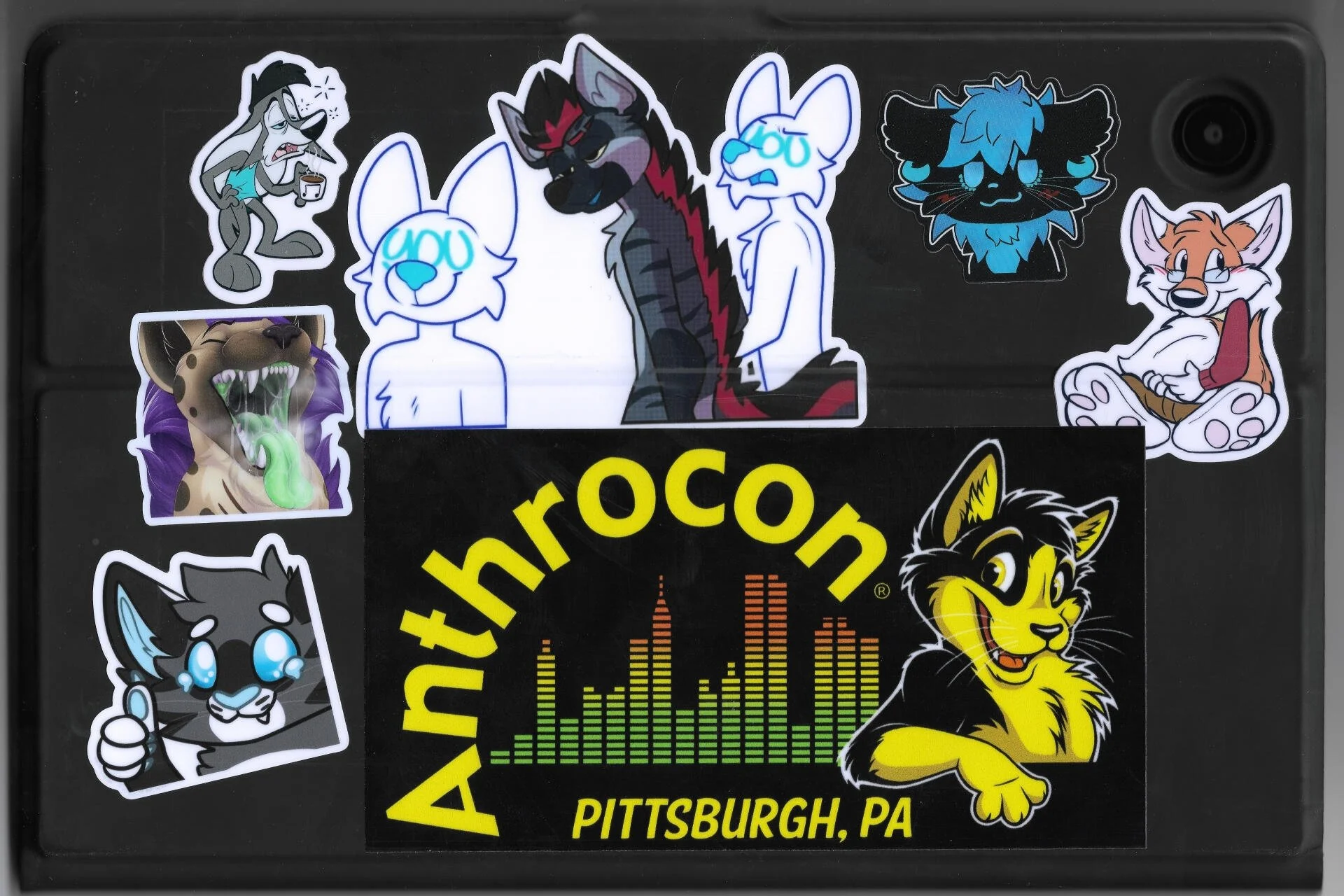 Collection of various colorful stickers featuring anthropomorphic animals, including a dog, cats, and a tiger, some with crying or angry expressions, and a large sticker with the text 'Anthrocon Pittsburgh, PA' with a yellow and black tiger mascot.