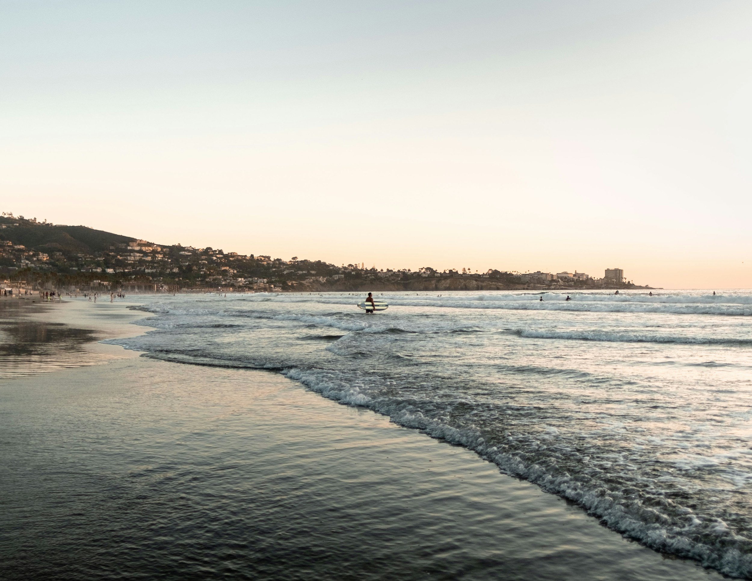 Sunset at the beach with waves and a person holding a surfboard in the water, shoreline with many people, hills and buildings in the background.