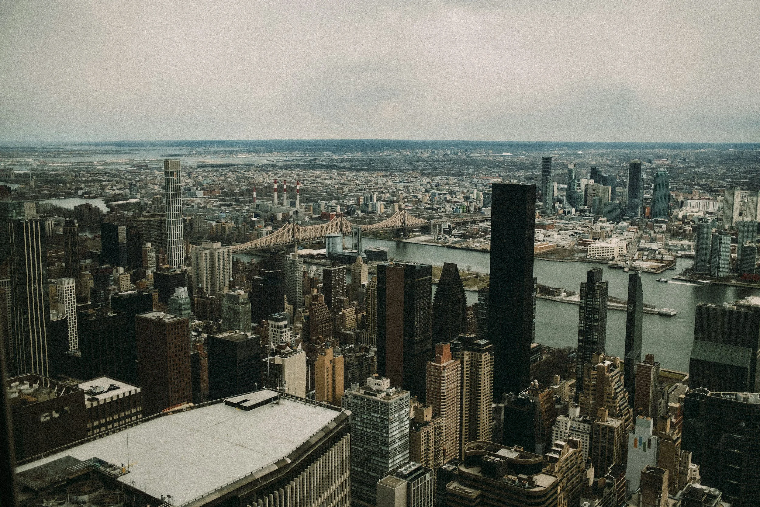 Aerial view of downtown New York City with skyscrapers, the East River, and the Queensboro Bridge under an overcast sky.