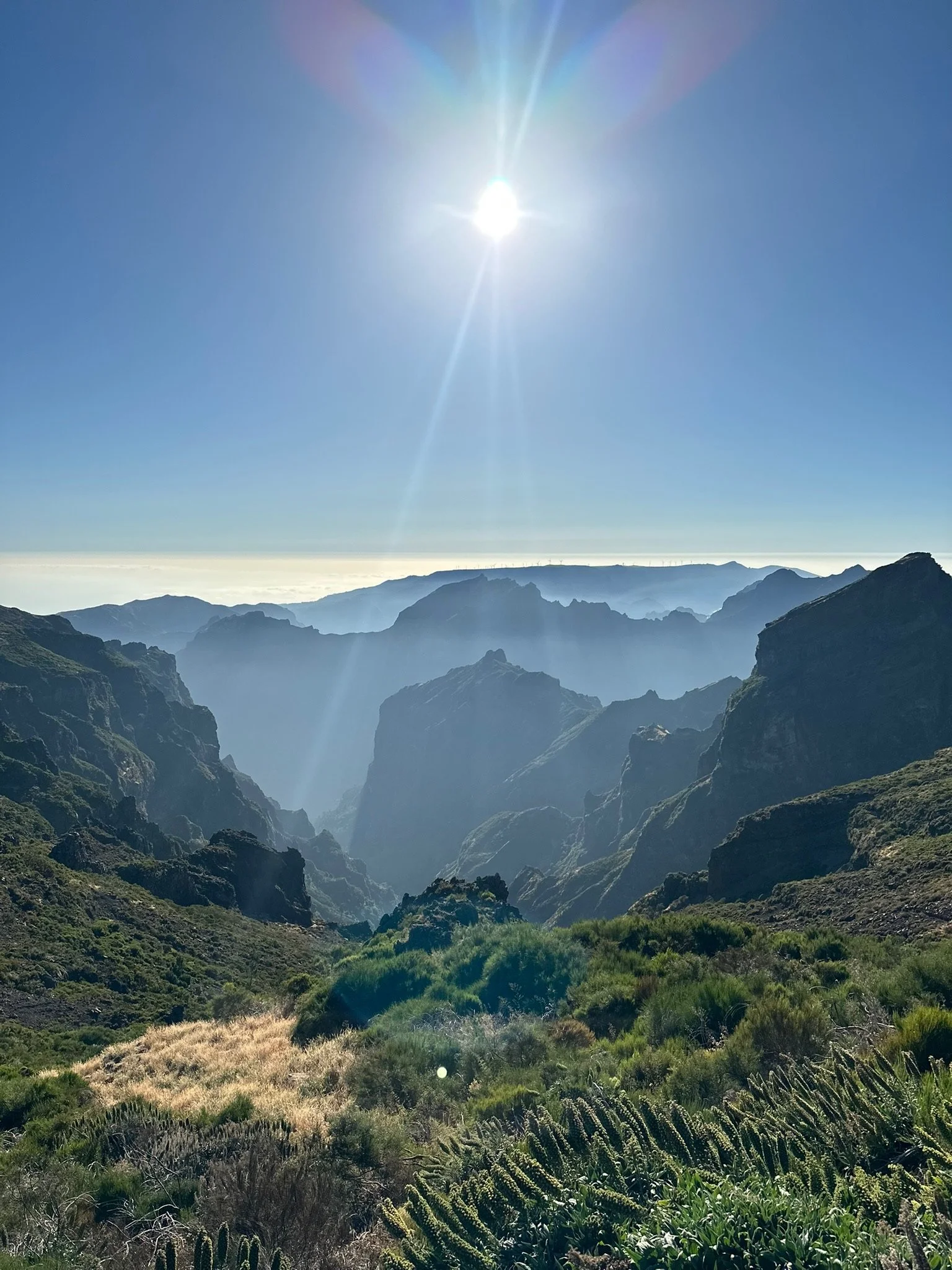 Sun shining over rugged mountain landscape with green vegetation in the foreground and multiple mountain ridges in the background under a clear blue sky.