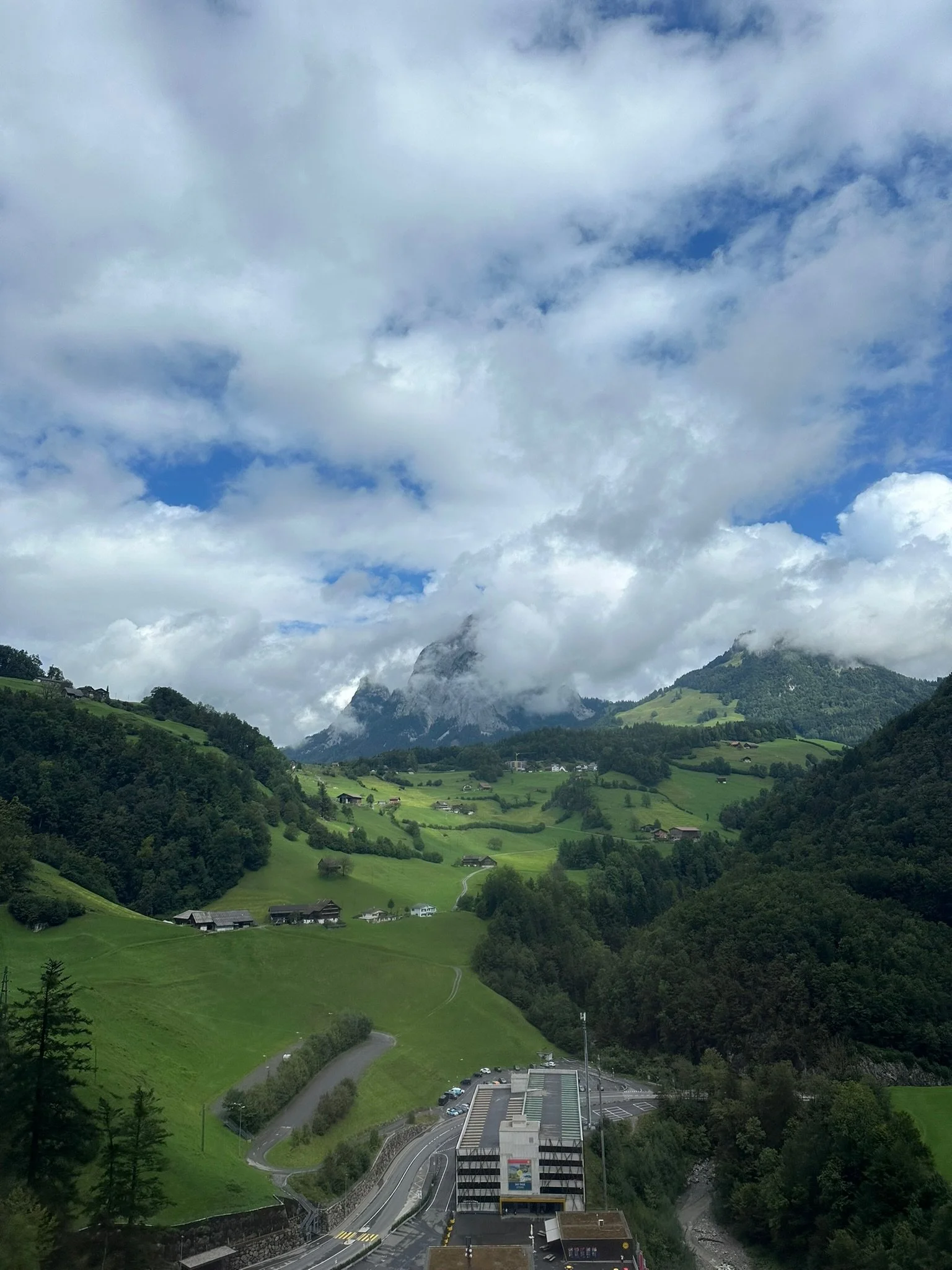 Scenic view of green rolling hills with scattered houses, a parking lot, and tall mountains partly covered by clouds in the background under a cloudy sky.