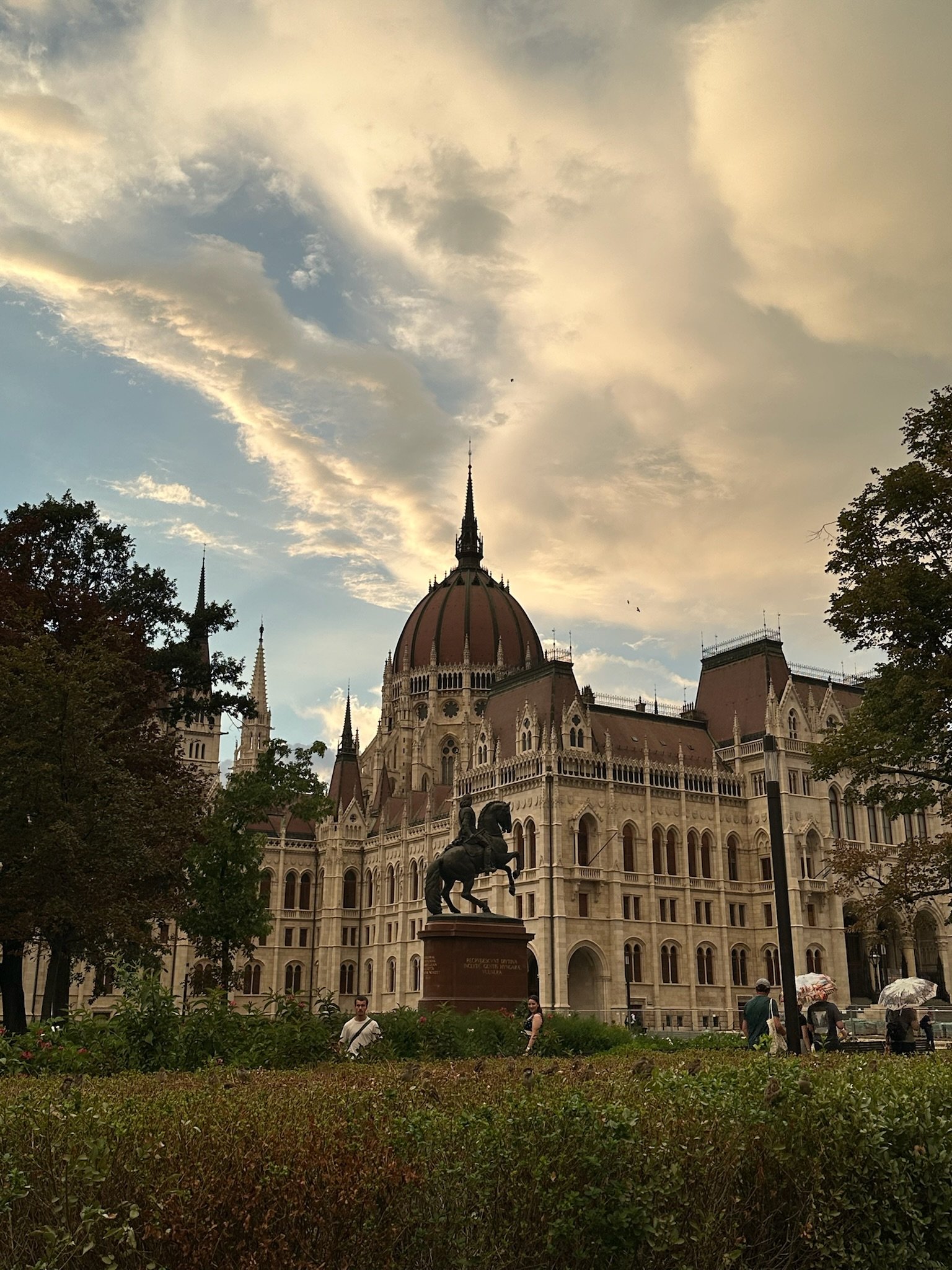 The Hungarian Parliament Building with a statue of a horse and rider in front, under a cloudy sky, and people walking nearby.