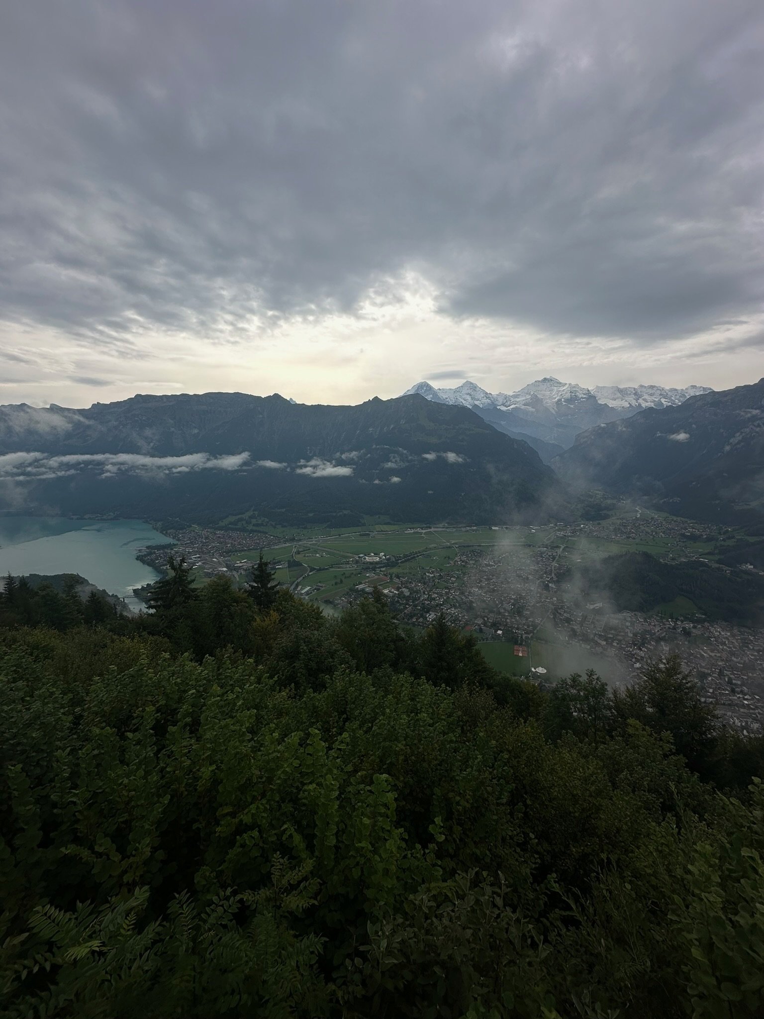 A mountain landscape with a cloudy sky, snow-capped peaks, a lake, and a town surrounded by green trees and fields in the foreground.