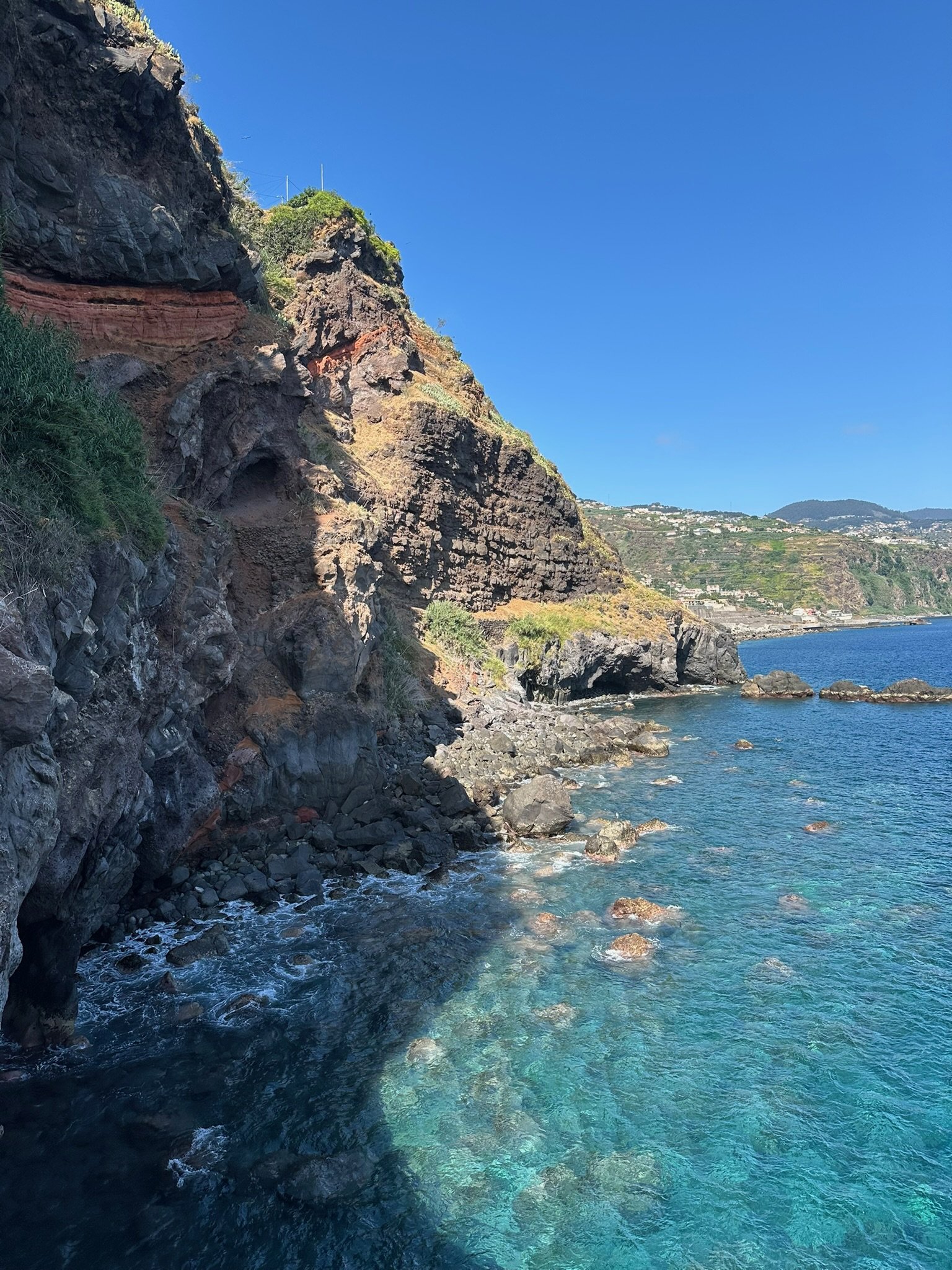 Coastal cliffs with rocky shoreline and clear blue water under a bright blue sky.