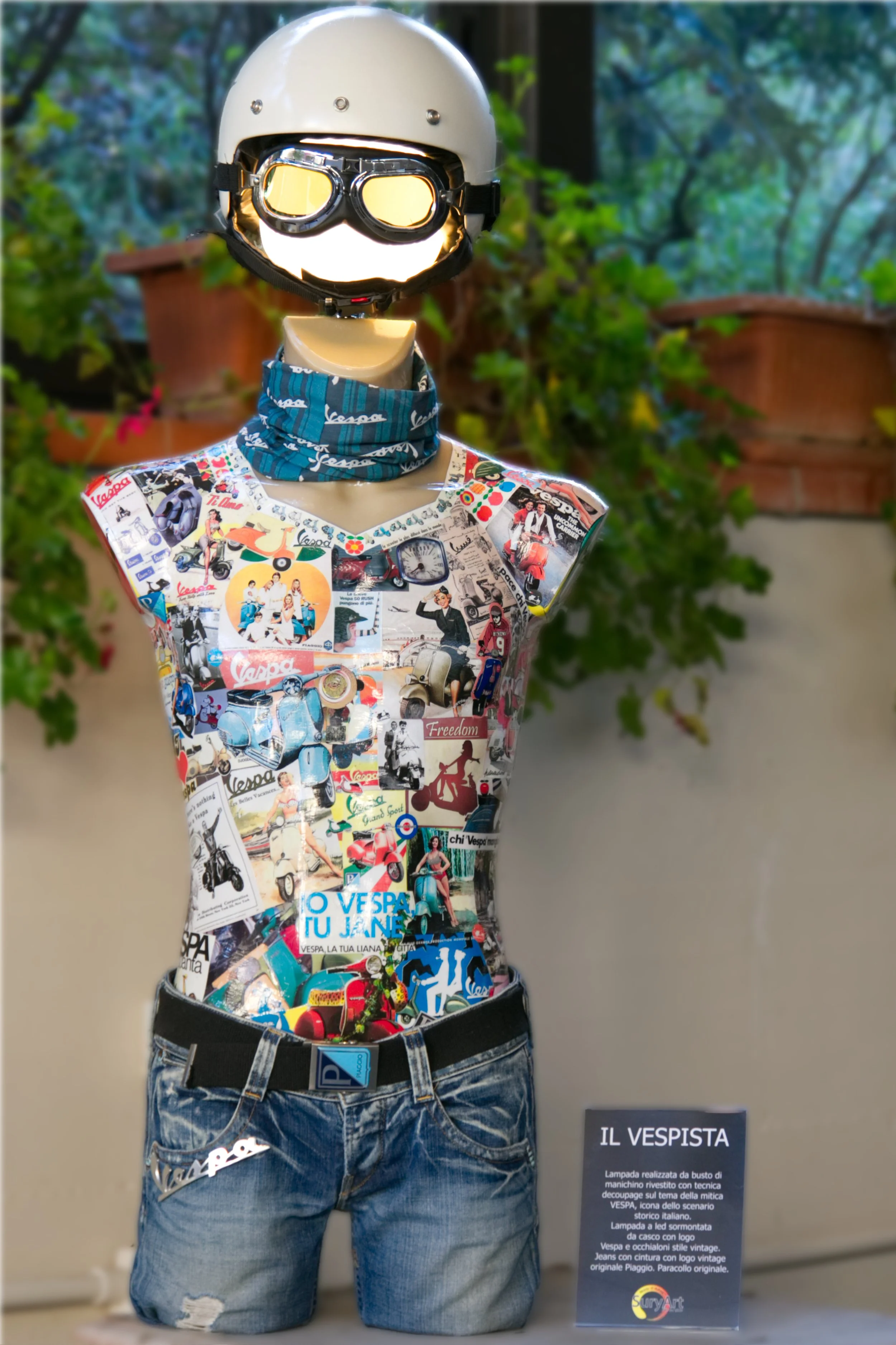 Scultura in stile vintage di un ragazzo con casco e occhiali da pilota, vestito con una maglietta a collage di pubblicità Vespa e jeans con tasca decorata e cintura originale Piaggio, esposta in una mostra con pannello informativo