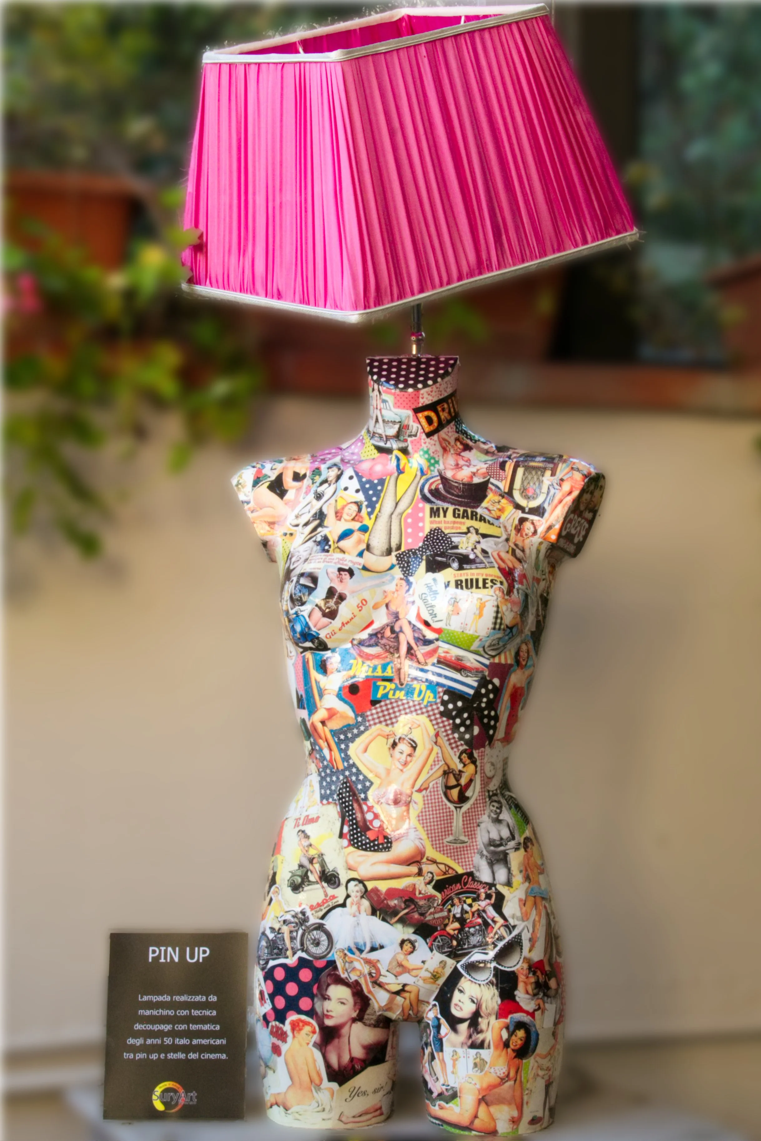 Statua di una donna decorata con collage di immagini vintage di pin-up, moda e cultura americana degli anni '50, con lampada rosa sopra. Un cartello a sinistra con scritto "PIN UP".