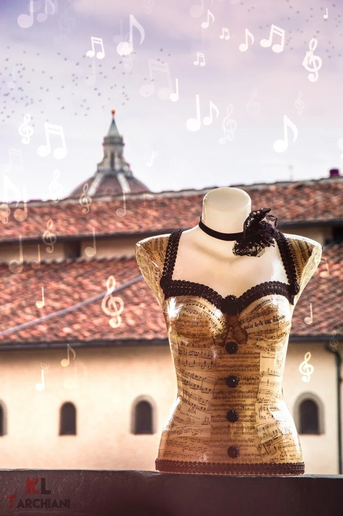 Una manichino vestito con un abito a forma di partitura musicale, con rifiniture nere, di fronte a un tetto di tegole rosse e un edificio con finestre ad arco, con sfondo un cielo con musica e note disegnate.