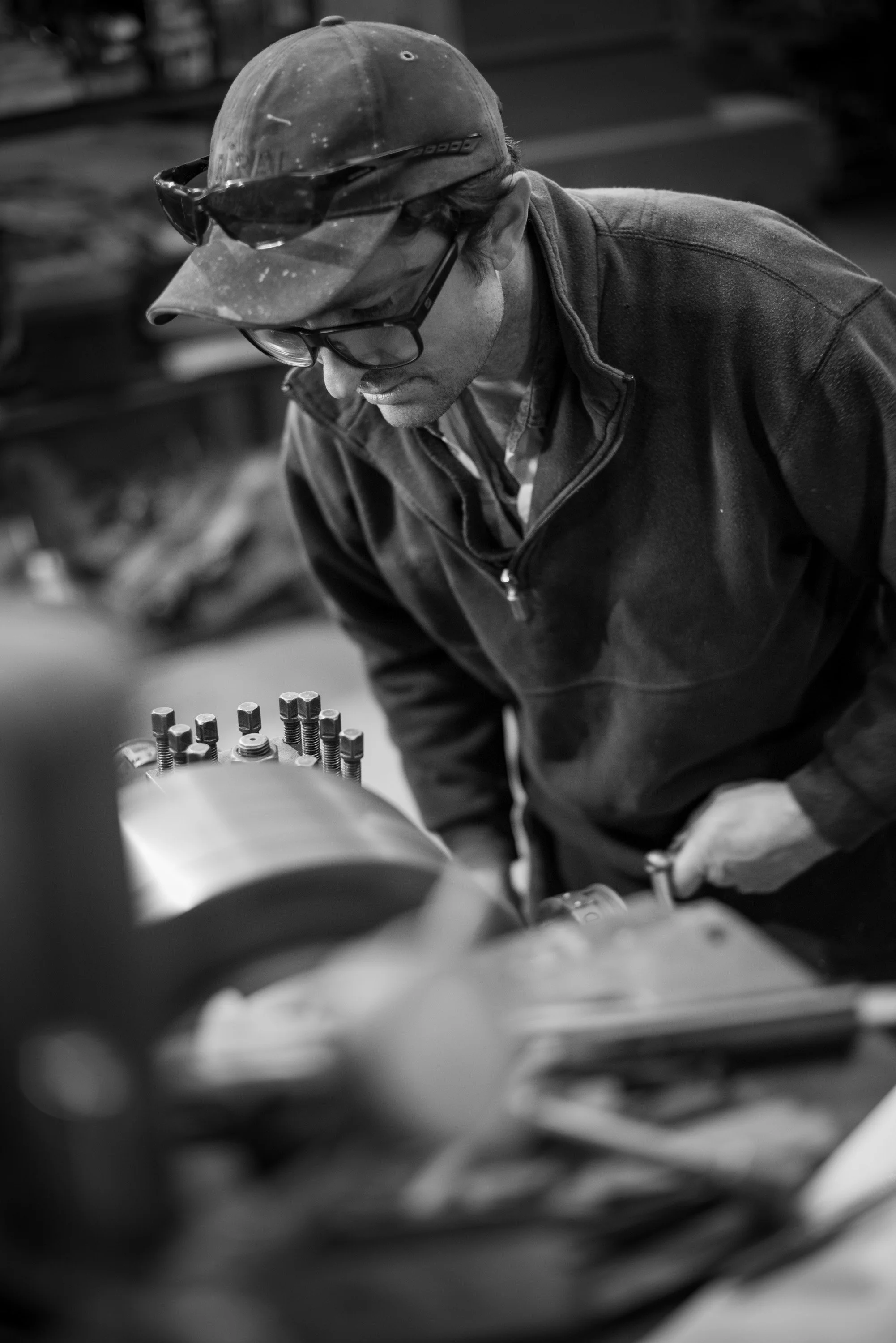 A man wearing glasses, a cap, and a jacket working on machinery in a workshop.
