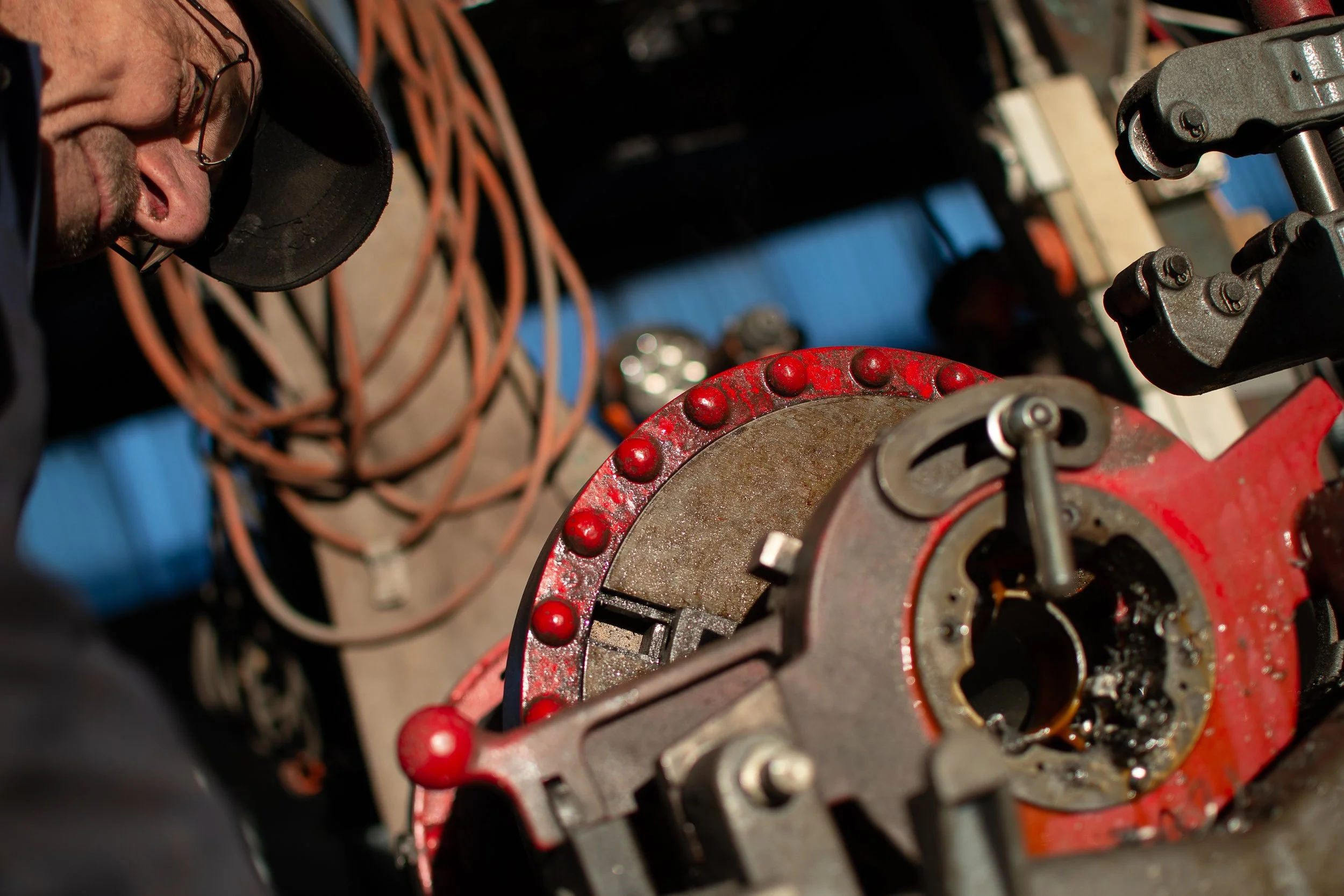A man working on a red mechanical part, possibly a large engine or machinery component, with his head tilted downward. Cables and tools are visible in the background.
