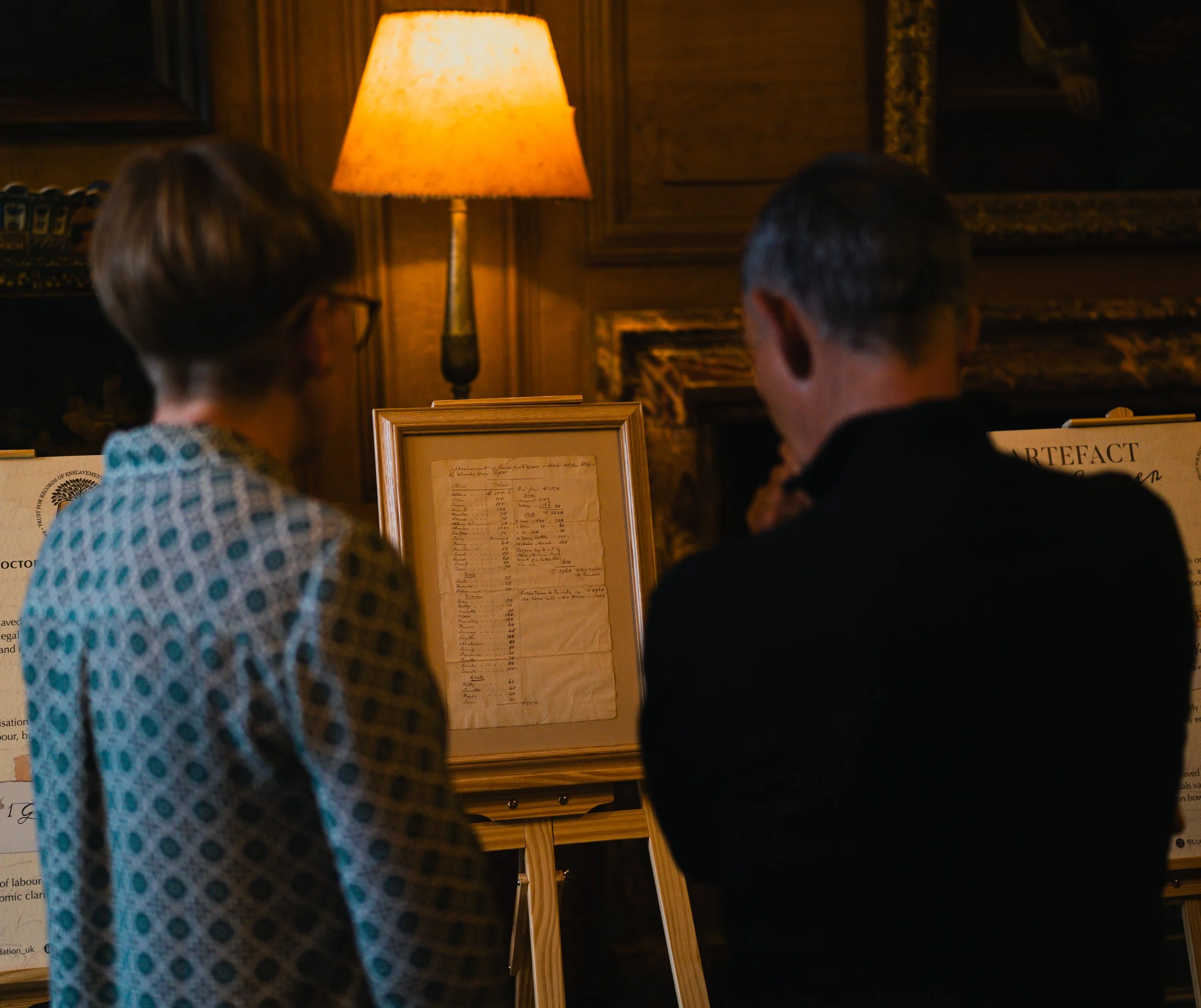Two people, one woman and one man, looking at a framed document displayed on an easel in a warmly lit room with wooden walls and a table lamp.