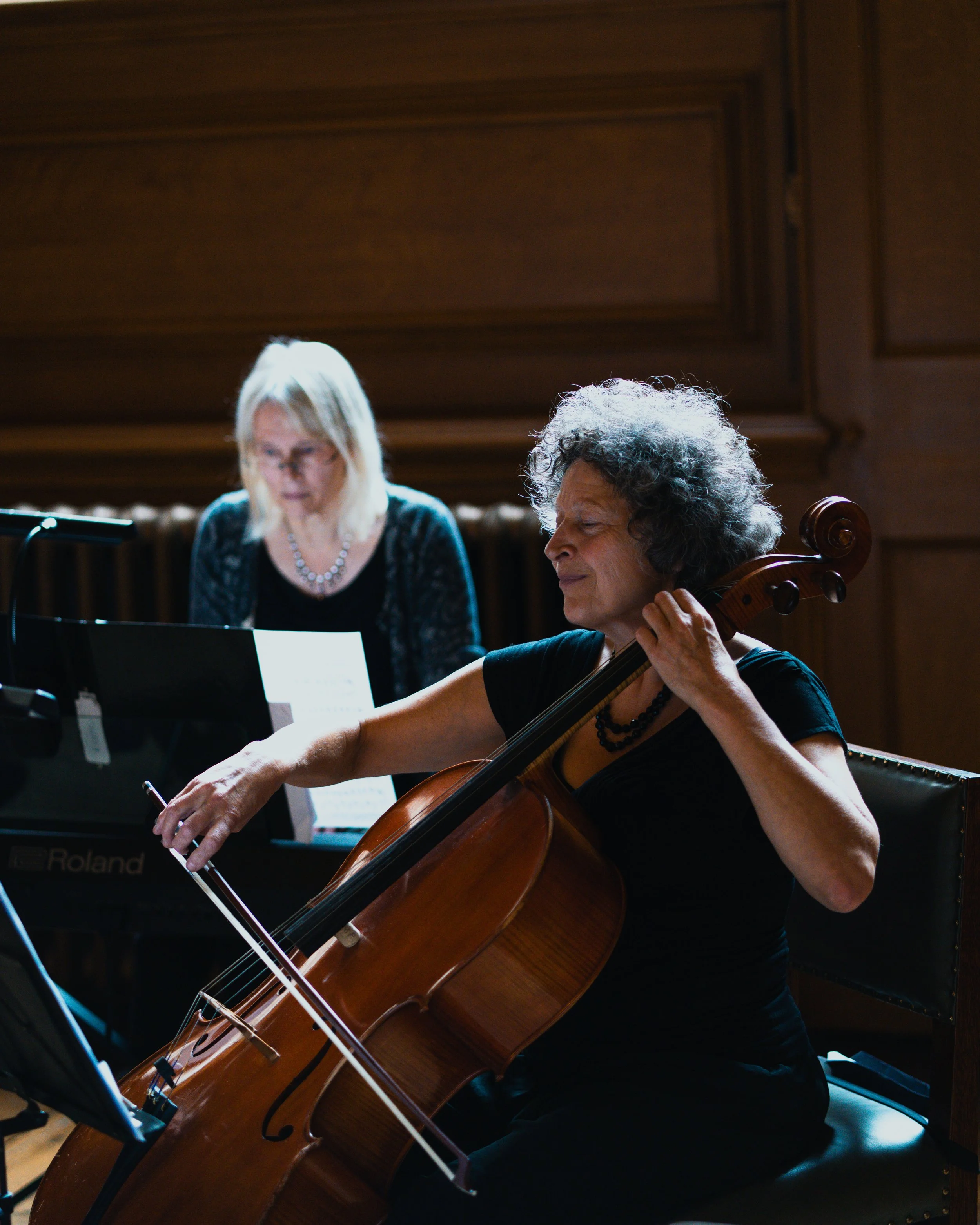 A woman playing the cello while two other musicians are in the background, one playing a keyboard.