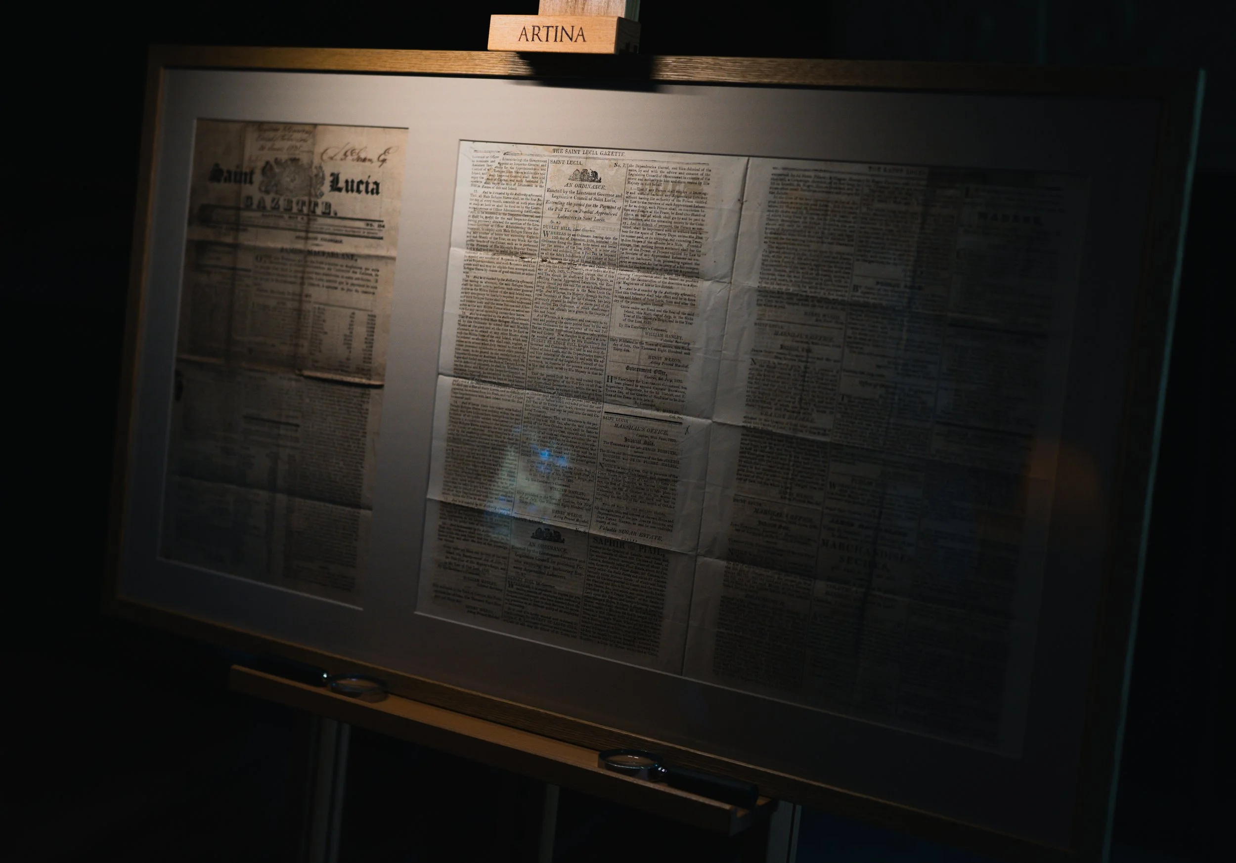 A framed display case containing an old newspaper with a sign labeled 'ARTINA' on top.