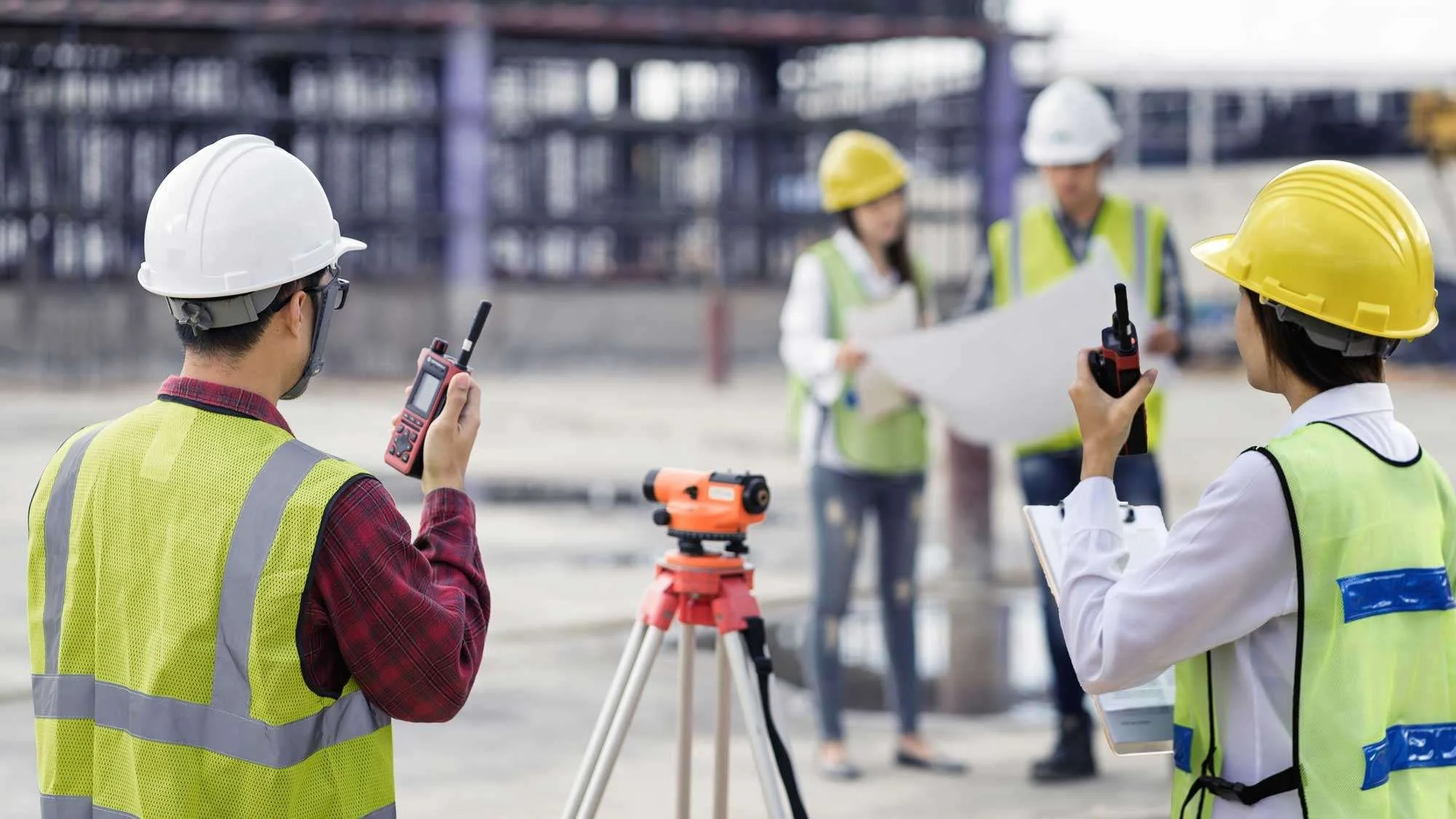 Construction workers and engineers on a site, wearing safety helmets and vests, using walkie-talkies and surveying equipment, with a worker holding blueprints.