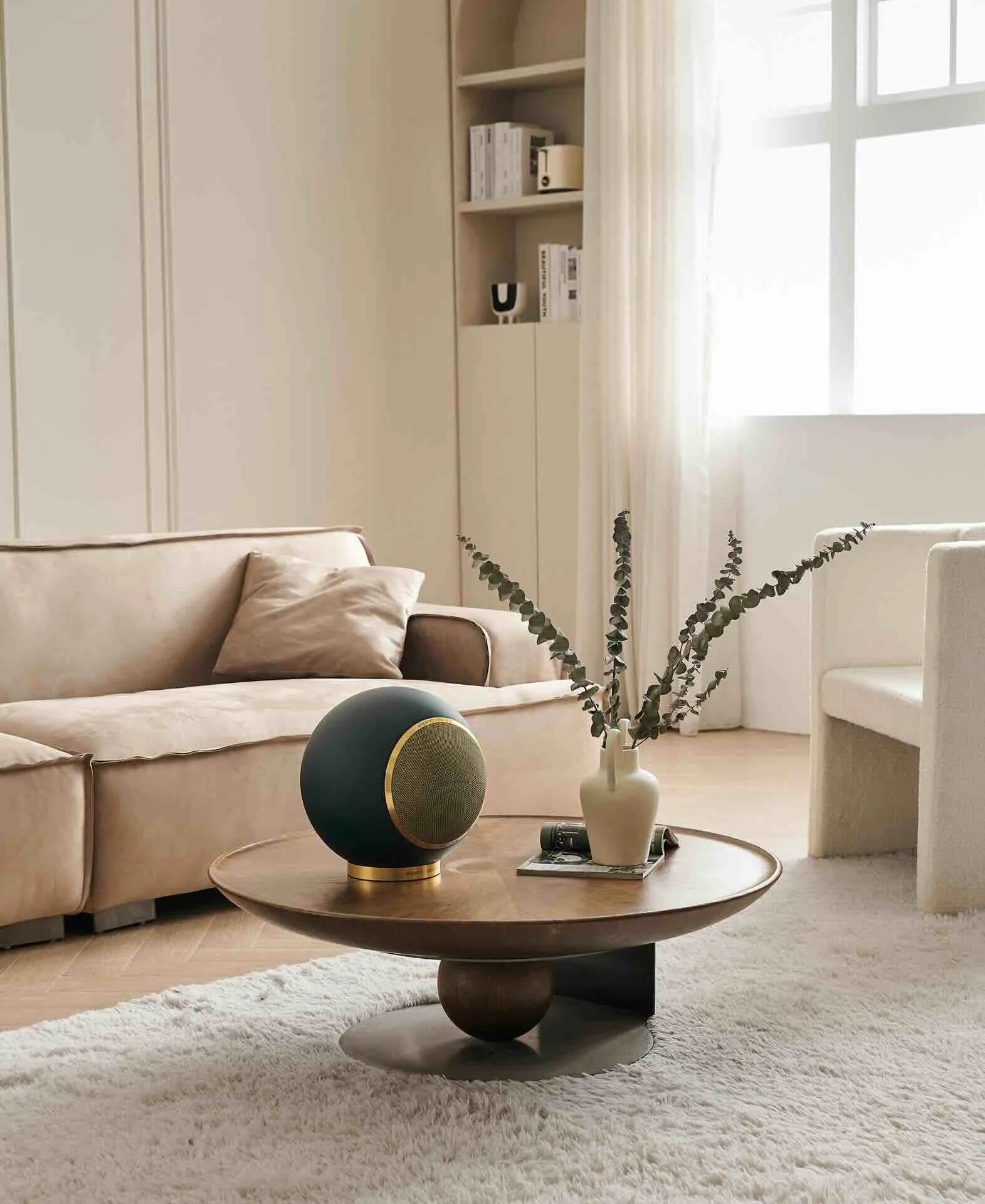 Ein modernes Wohnzimmer mit einem beigen Sofa, einem Teppich und einem niedrigen Couchtisch, auf dem sich eine weiße Vase mit Zweigen, eine schwarze runde Lautsprecher und Zeitschriften befinden. Im Hintergrund sind ein Regal und ein Fenster mit weißen Vorhängen.
