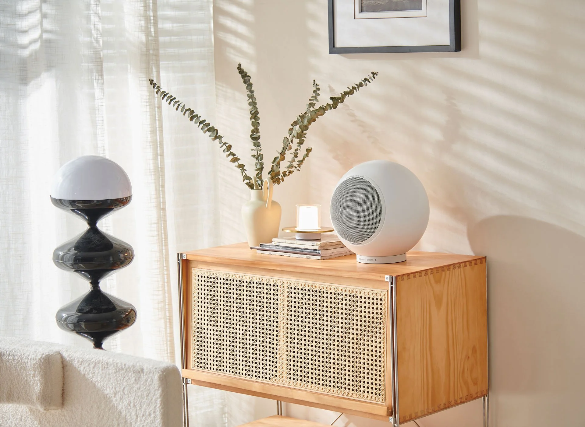 Ein Holzsideboard mit einer weißen runden Lautsprecher, einer weißen Vase mit langen grünen Zweigen, einem kleineren Licht und einigen Büchern darauf. Im Hintergrund eine weiße Wand und weiße Vorhänge.