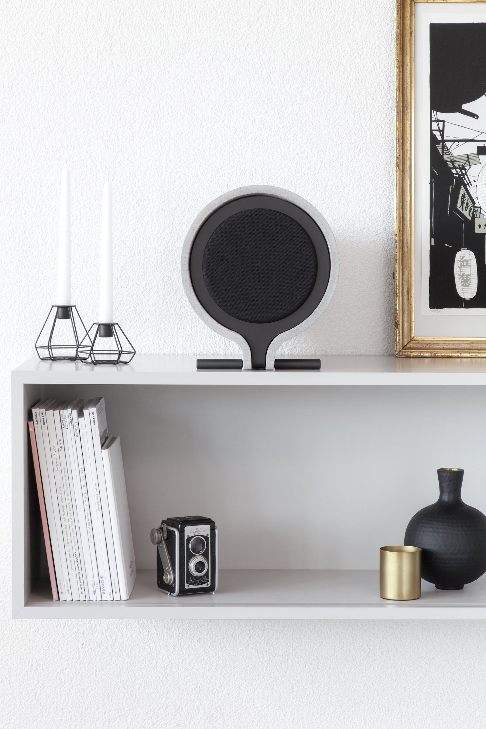 Weiße Wand mit einem weißen Regal, darauf ein moderner Lautsprecher, zwei weiße Kerzen in schwarzen Metallhaltern, eine analoge Kamera, schwarze und goldene Dekorvasen, eine Wandkarte mit japanischen Schriftzeichen.