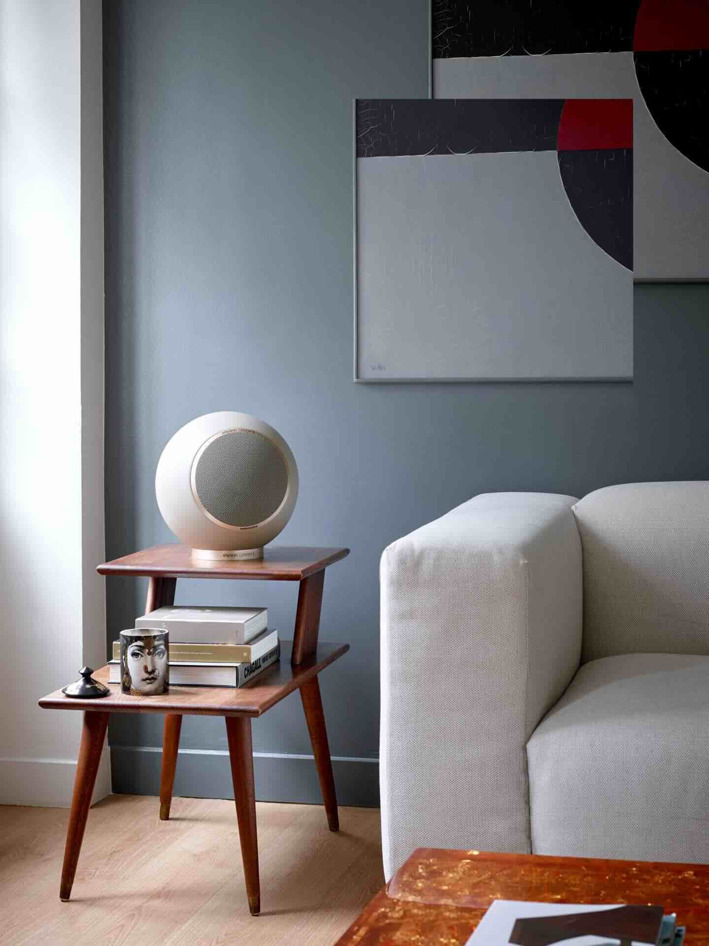 Ein Wohnzimmer mit einem weißen Sofa, einem kleineren Beistelltisch aus Holz, auf dem ein Lautsprecher, ein Buch, eine Kerze und eine kleine Figur stehen. An der Wand hängen abstrakte Kunstwerke.