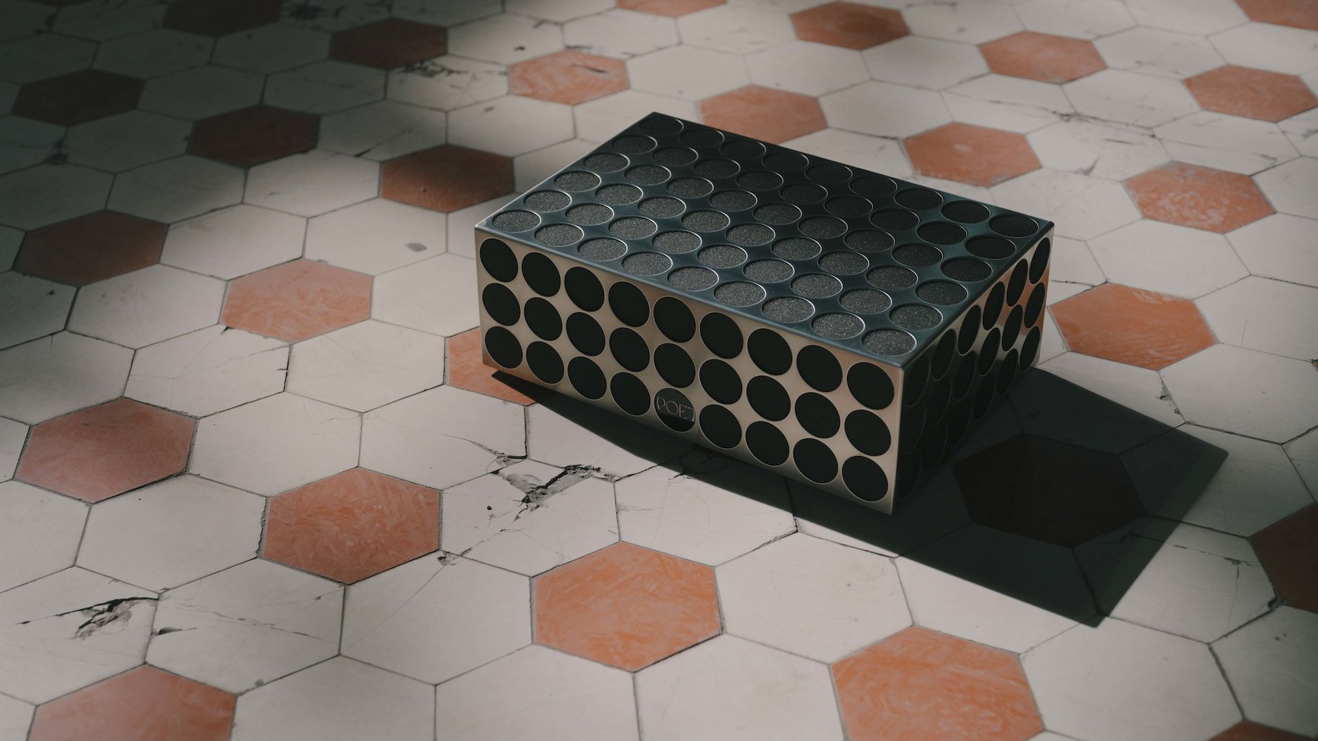 Ein Edelstahl Soundsystem mit runden verstiefungen im Gehäuse liegt auf einem verfliesten Boden mit weißen und roten Fliesen.