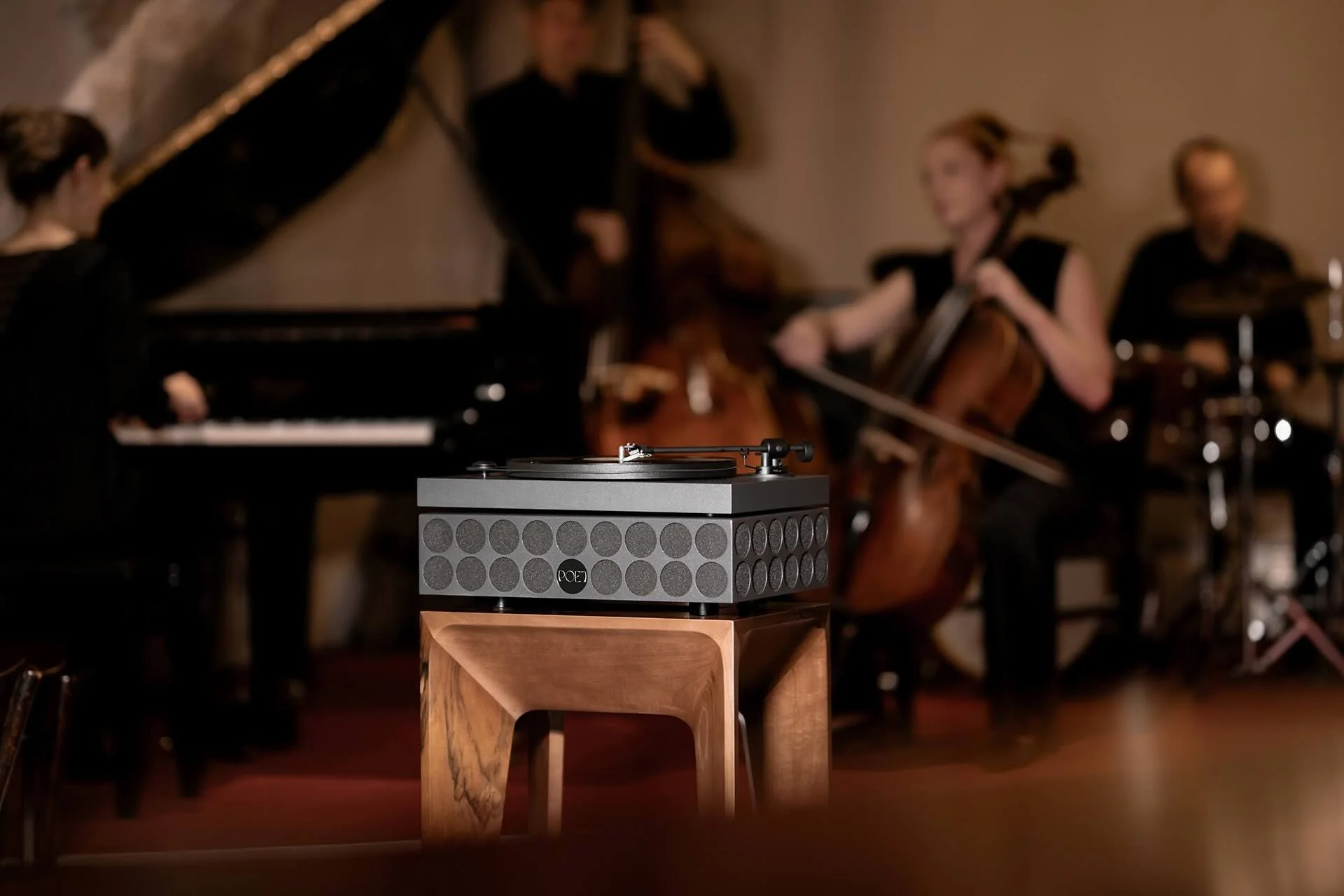 Eine POET fonetta° steht auf einer base° aus Holz, direkt neben einem Klavier.
Im Hintergrund spielt eine Jazzband mit Kontrabass, Cello, Schlagzeug und Piano.
Die Szene wirkt warm, intim und musikalisch — ein Zusammenspiel aus akustischen Instrument