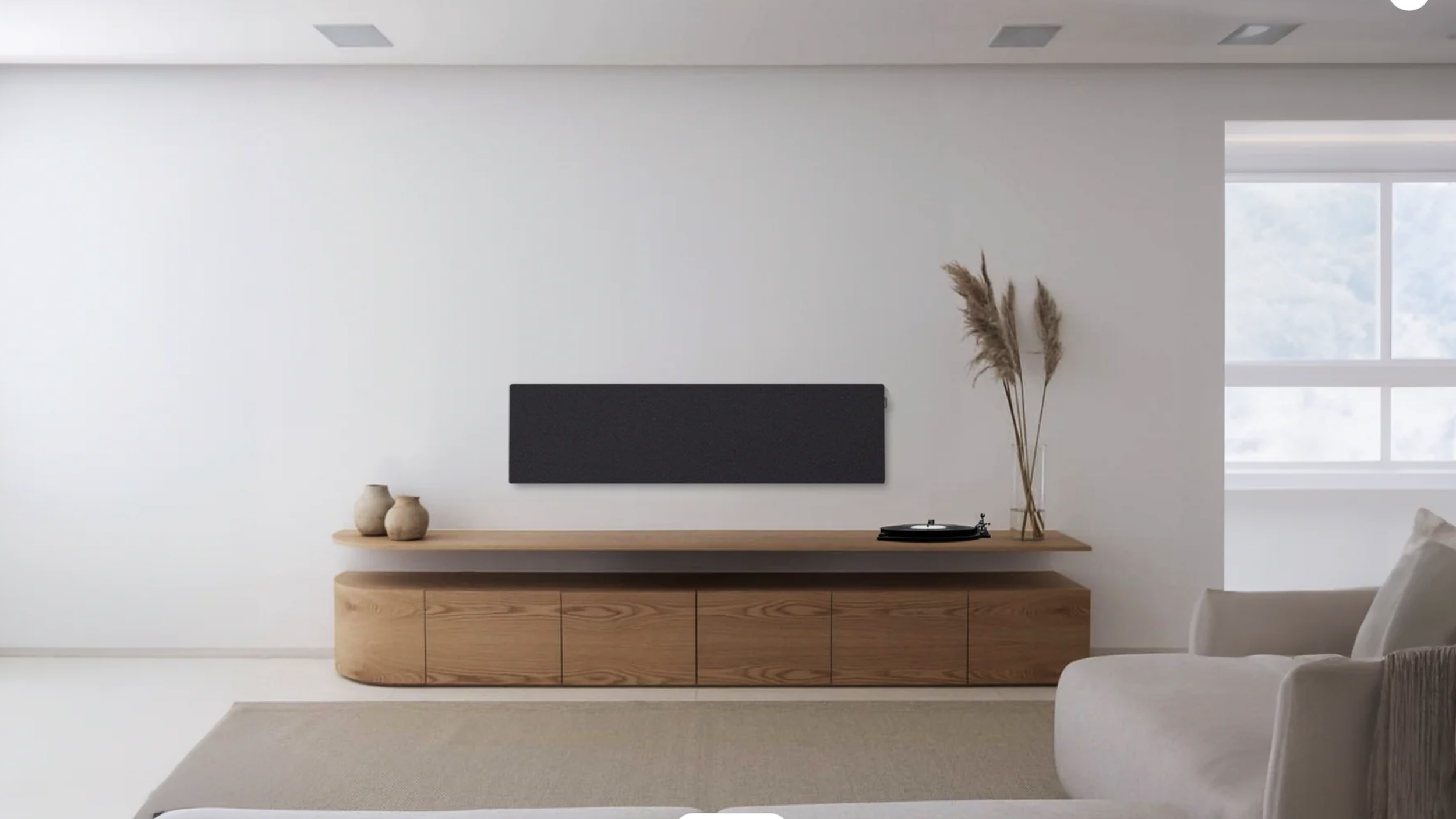 Ein minimalistisches Wohnzimmer mit einem weißen Sofa, einem langen hölzernen Lowboard, auf dem drei Keramikvasen, ein Plattenspieler und ein Vase mit getrockneten Gräsern stehen.