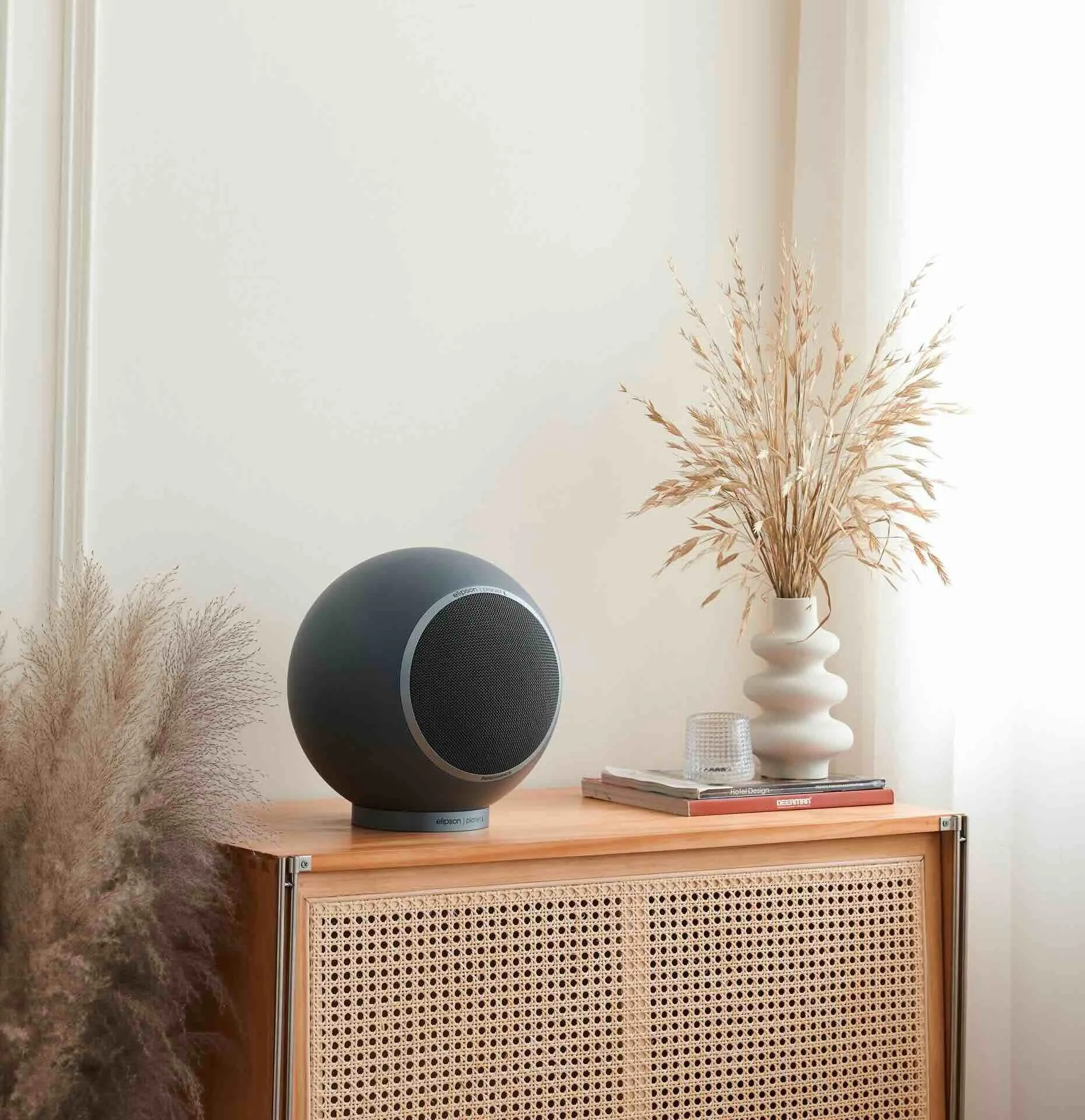 Ein moderner, runder Lautsprecher auf einem Holzsideboard neben einem stilvollen Vase mit getrockneten Gräsern und einem Glas, bei Tageslicht.
