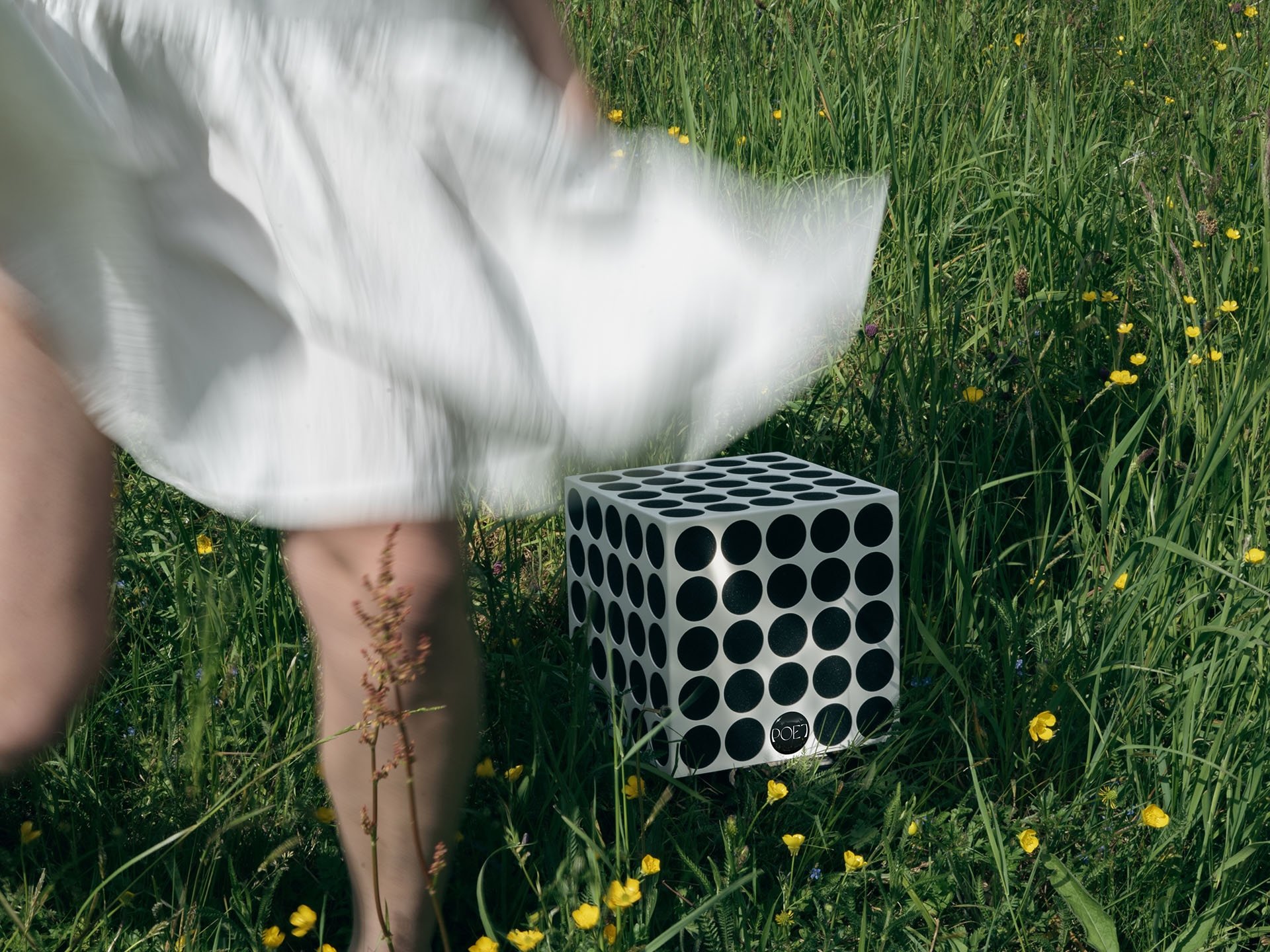 Eine Frau in einem weißen Kleid steht bzw. bewegt sich auf einer Wiese mit hohem Gras und Wildblumen.
Vor ihr steht eine weiße POET zero°, deren charakteristische Punktstruktur klar erkennbar ist.
Die Szene wirkt leicht, natürlich und sommerlich – ei