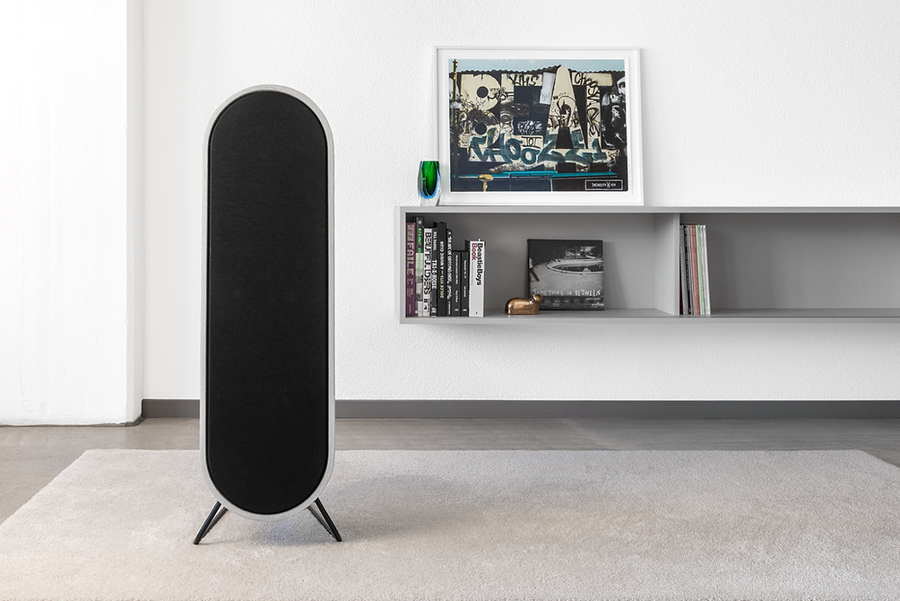 Moderner schwarzer Lautsprecher in einem minimalistisch eingerichteten Raum, vor weißer Wand mit Regal und Kunstwerk.