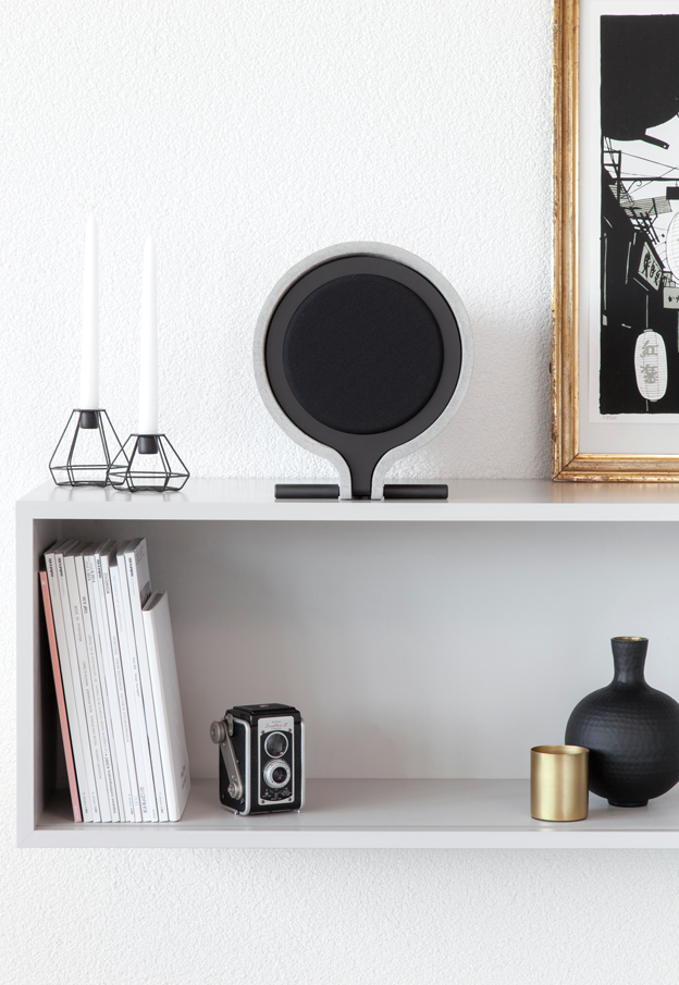 Weiße Wand mit weißem Regal, auf dem stehen zwei Kerzen in geometrischen Haltern, eine Vintage-Kamera, schwarze Vase, goldene Kerze, schwarze Platte, und auf dem Regal befinden sich auch Bücher und ein Lautsprecher.