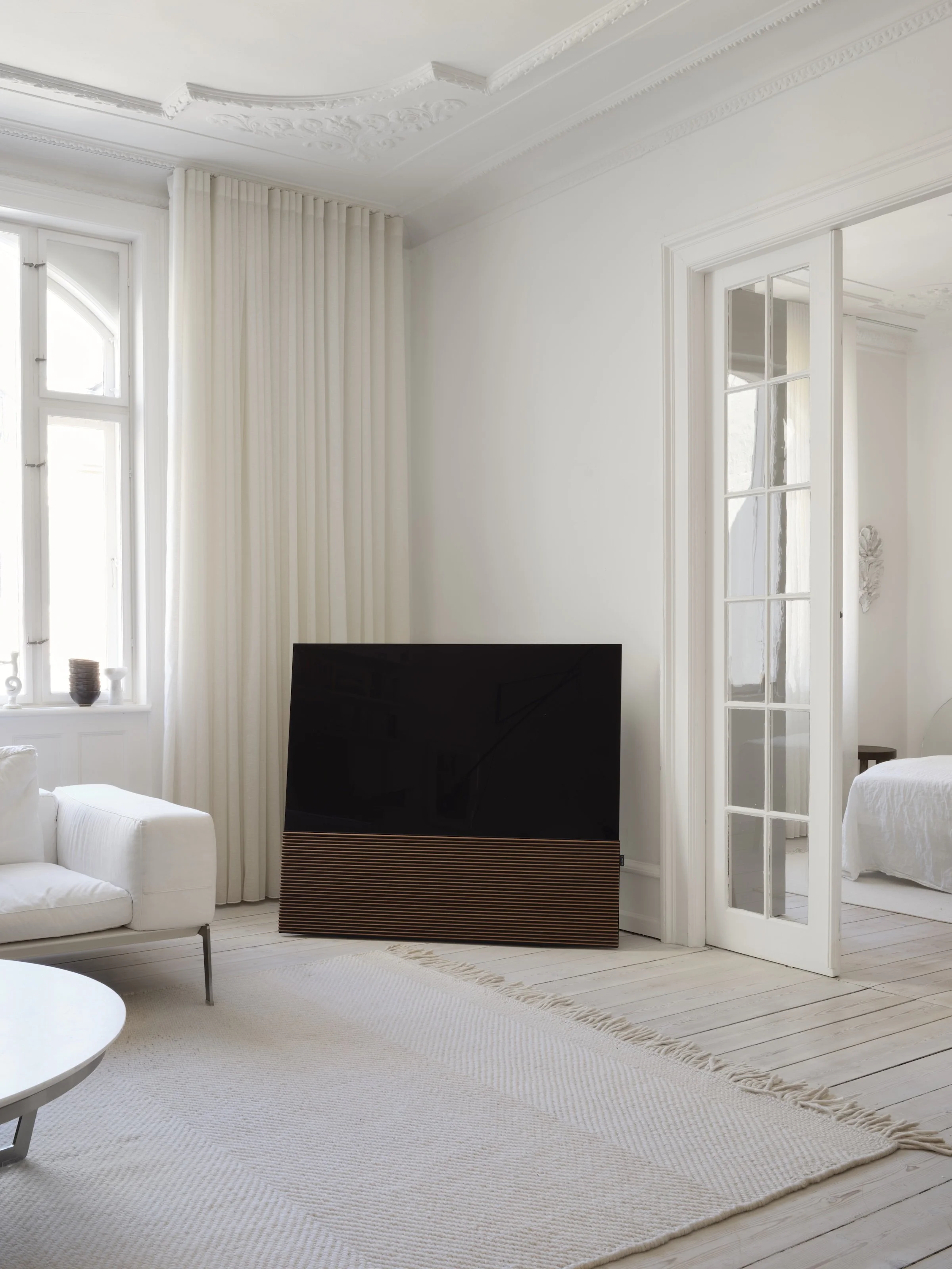 Ein weißes Wohnzimmer mit großen Fenstern, weißen Wänden, einem schwarzen Fernseher und einer weißen Couch.