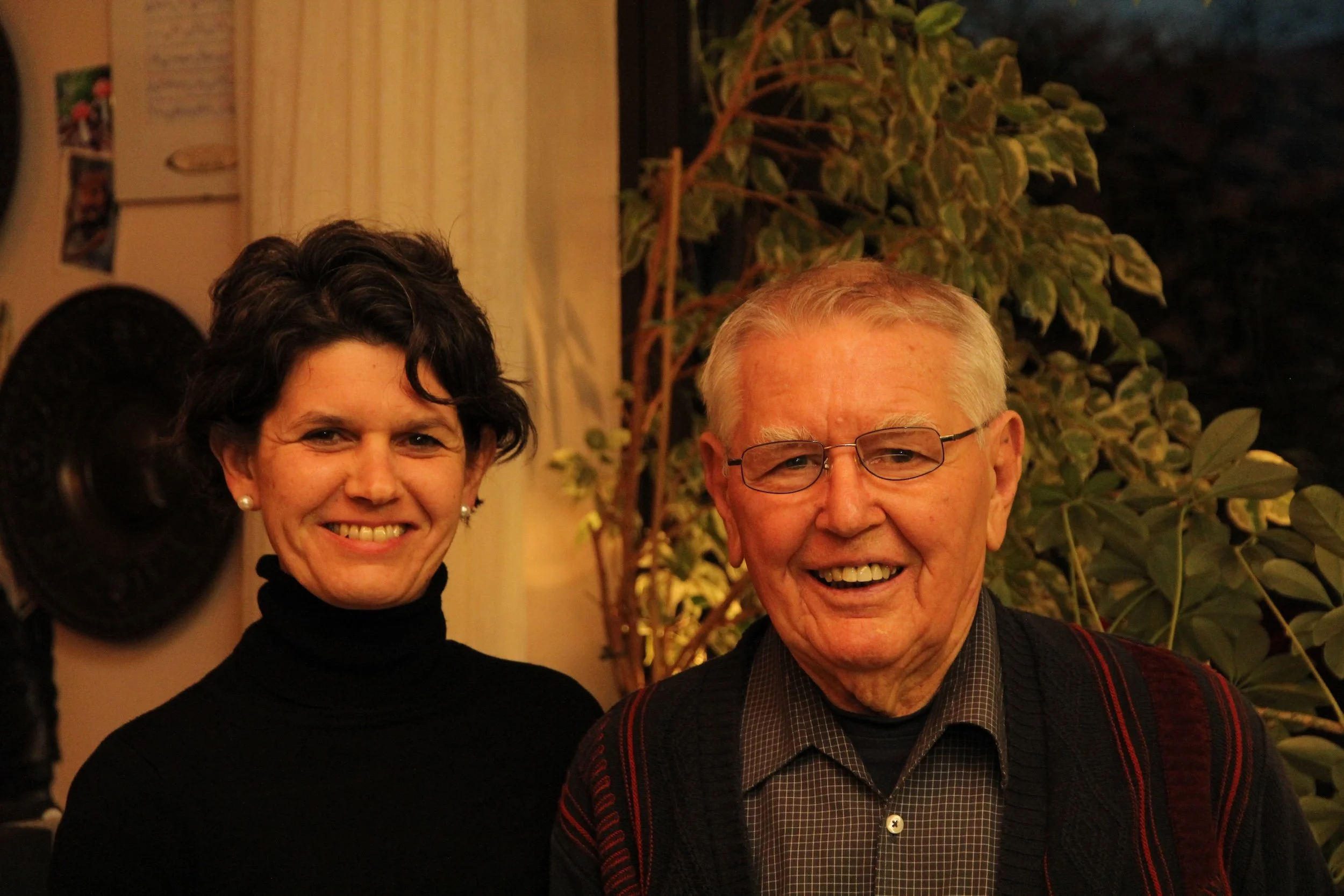 Zwei lachende Menschen, eine Frau und ein Mann, stehen nebeneinander vor einem grünen, blattbedeckten Hintergrund, in einem angenehmen, warm beleuchteten Raum.