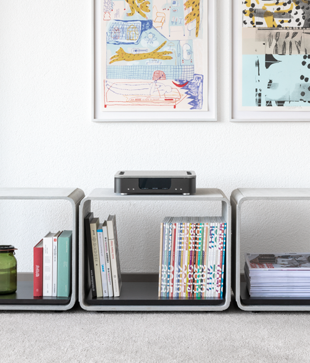 Regal mit Büchern, ein Drucker auf einem kleinen Tisch, zwei Regale an den Seiten, und Kunstwerke an der weißen Wand