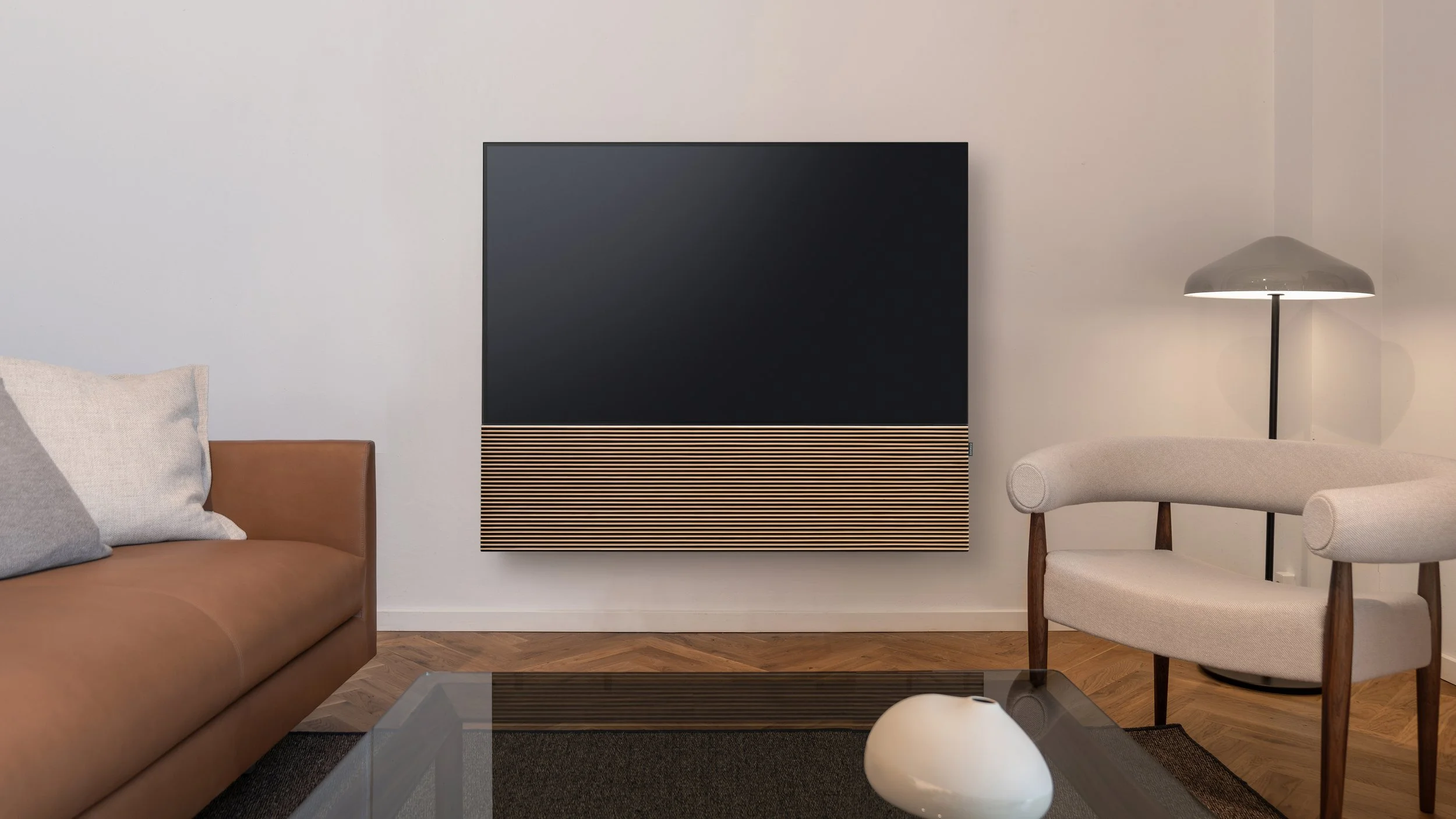 Modernes Wohnzimmer mit braunem Ledersofa, weißen Kissen, einem schwarzen Fernseher mit Holzelementen, einem weißen Sessel mit Holzbeinen, einem schwarzen Stehlampe und einem Glastisch mit einer weißen Vase.