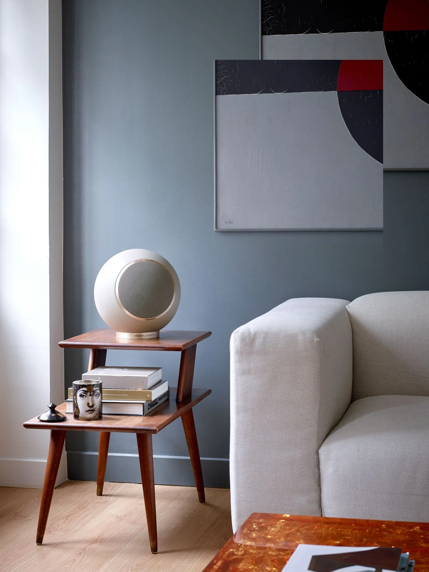 Ein Wohnzimmer mit einem Sofa, einem Holztisch, einem Buch, einem Kunstwerk an der Wand und einem modernen Lautsprecher auf dem Tisch.