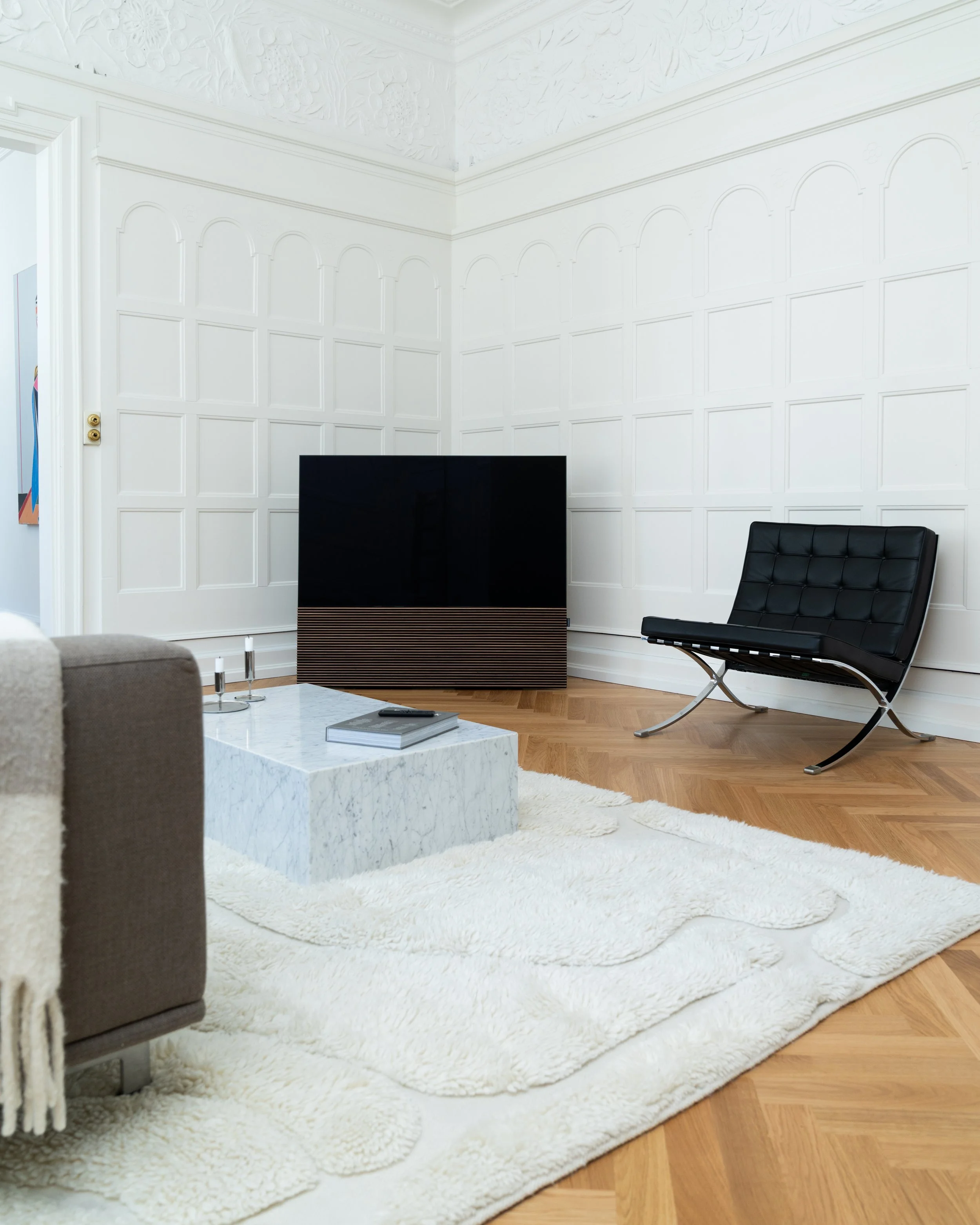 Modernes Wohnzimmer mit weiß verfugten Wänden, Holzparkett und hellen Teppichen. Es gibt einen schwarzen Fernseher, einen schwarzen Lounger und einen weißen Marmortisch.